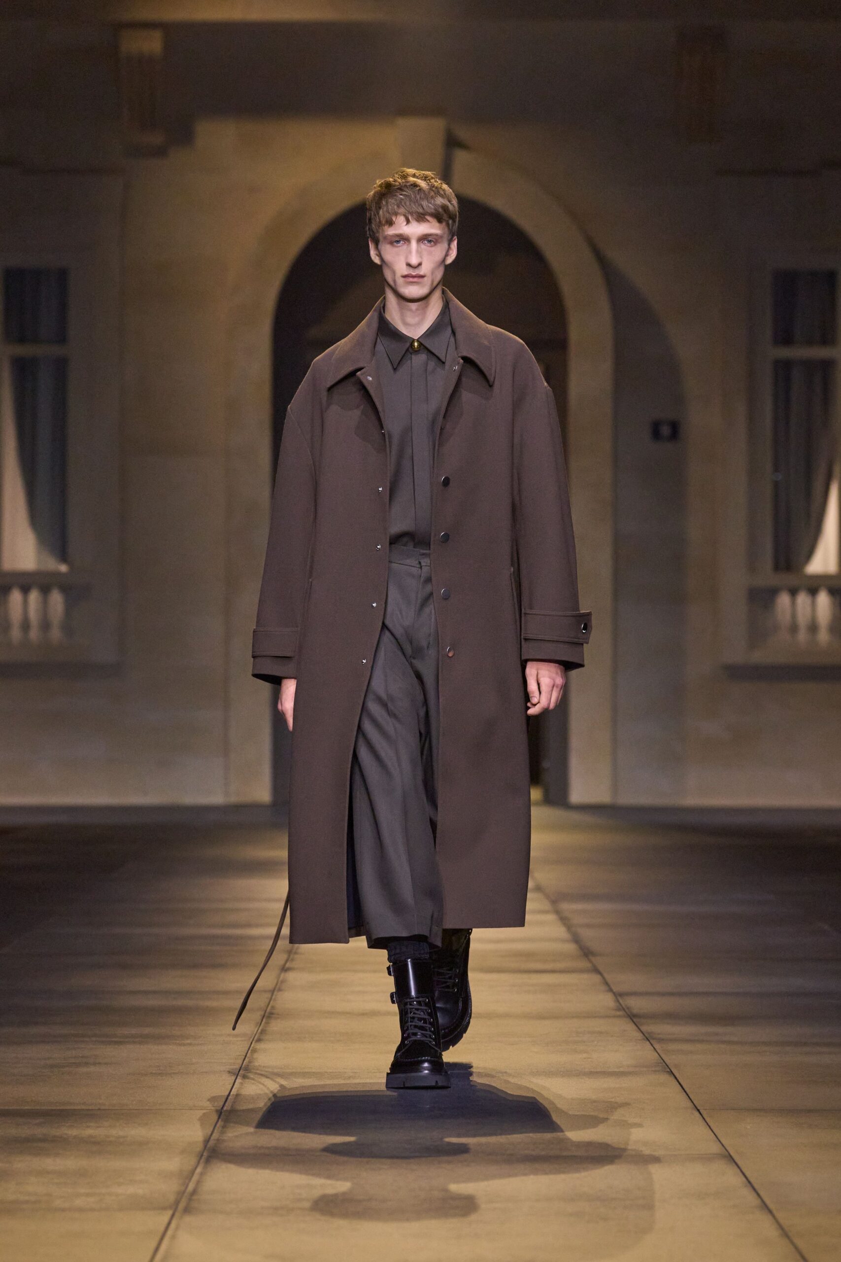 Dark cocoa overcoat with snap front and a belt end dragging. Smoke-grey shirt pinned shut with a single gold collar stud. Matching trousers with a soft pressed crease. Black lace-up combat boots with side buckles. Crossing a stone courtyard before the offices open.