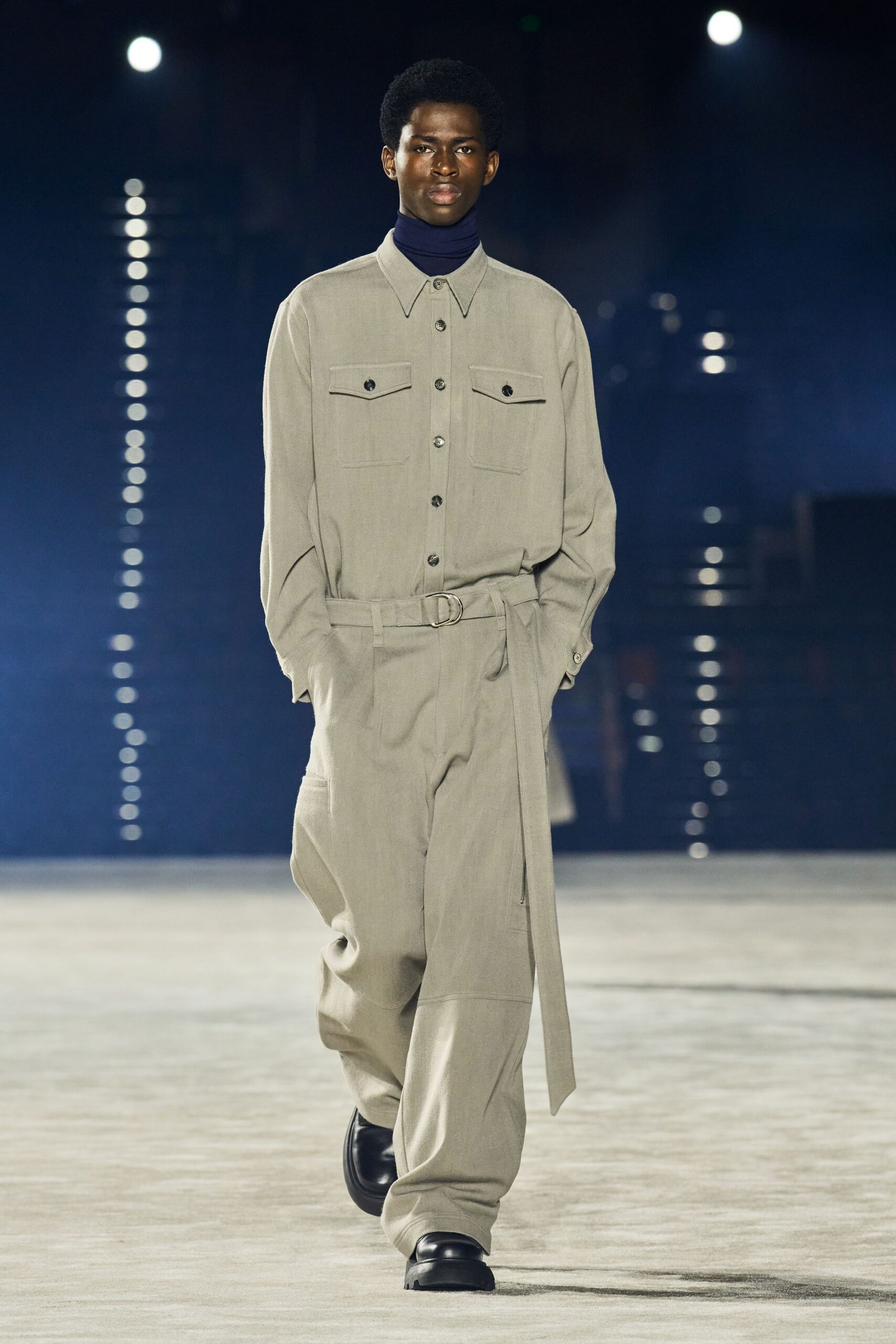 Ink-navy fine-knit turtleneck tight to the throat. Stone twill jumpsuit with pointed collar and flap chest pockets. Self belt threaded through a silver D-ring, tail hanging long. Black rounded shoes with thick lug soles. Walking across an empty ferry terminal before the first announcement.
