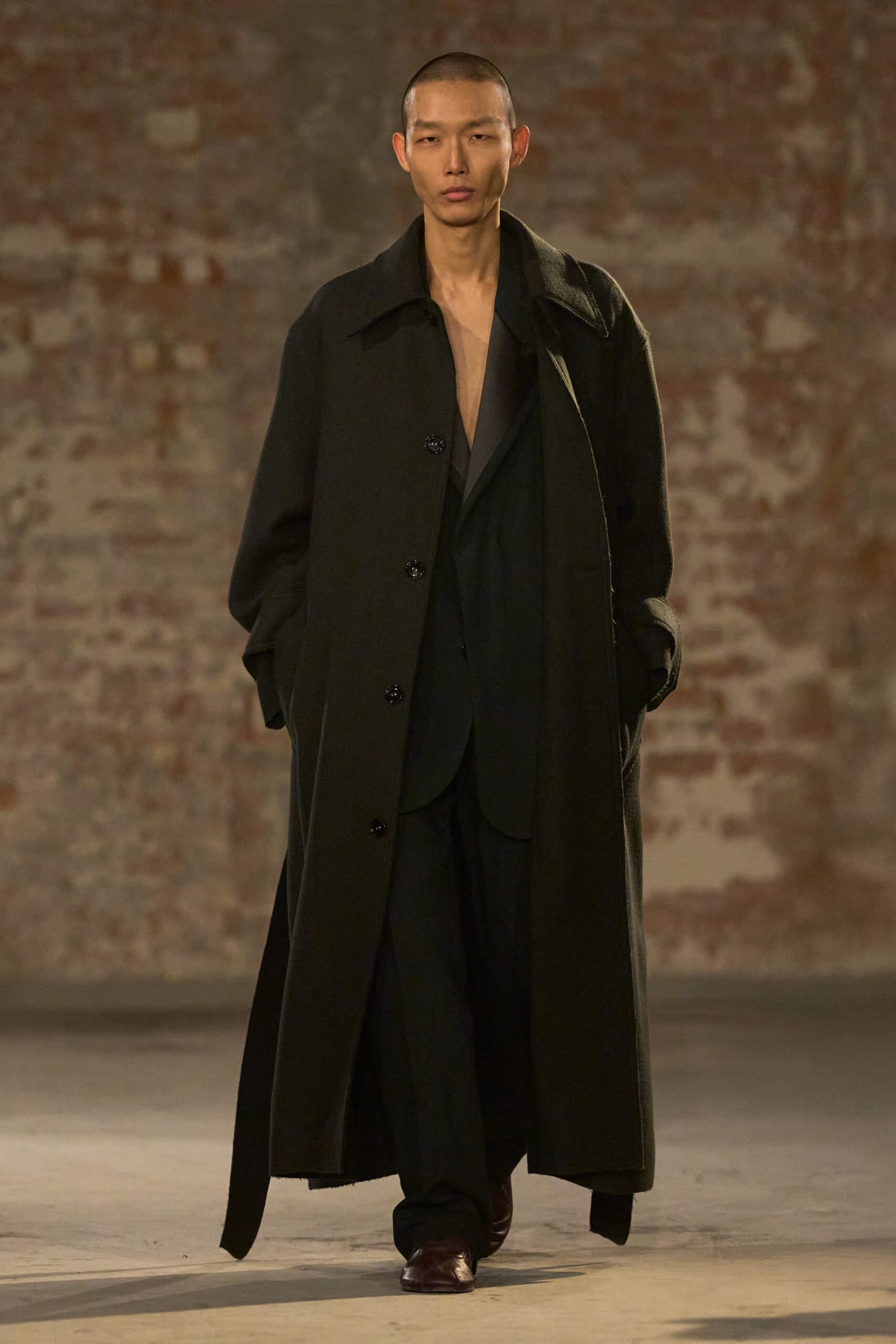 Long black wool coat with broad collar and glossy horn buttons. Black blazer hanging open, lapels dipping into a bare V. Loose black trousers breaking softly over the shoe. Oxblood square-toe leather shoes catching the light at the vamp. Crossing a drafty station hall after missing the last daylight train.