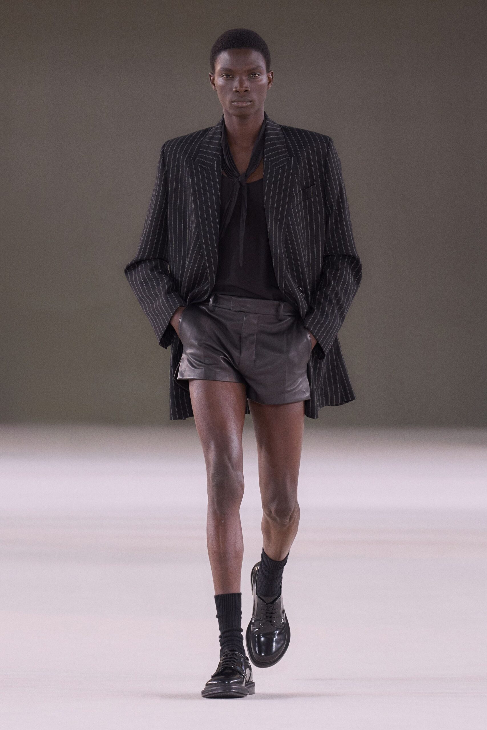 Long charcoal pinstripe blazer with sharp shoulders and a slanted welt chest pocket. Sheer black blouse tied loosely at the throat. Black leather shorts with a pressed front crease. Ribbed black crew socks. Glossy black derbies with a squared apron toe. For crossing the hotel lobby before breakfast in yesterday's clothes.