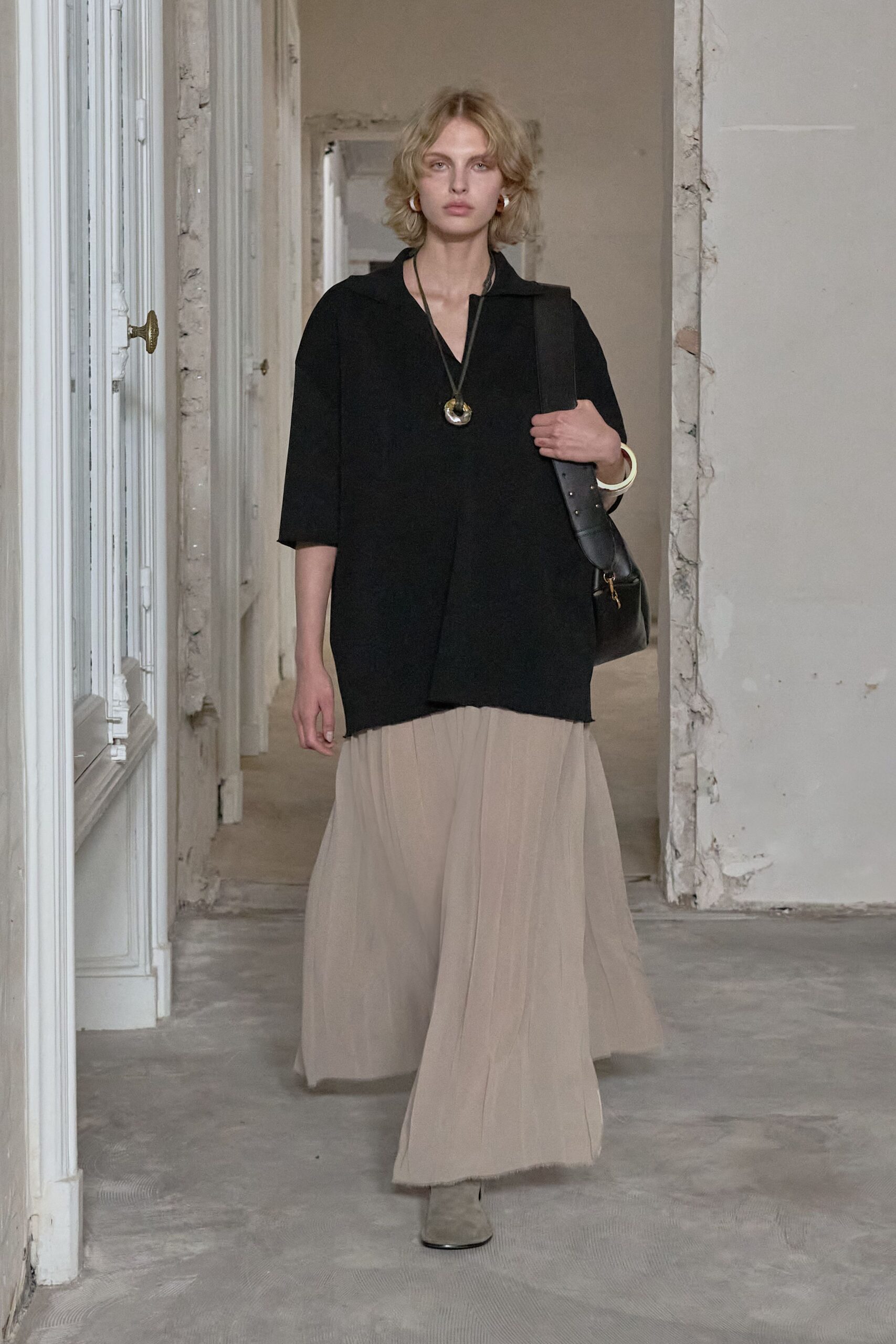 Black V-neck tunic, raw hem dipping lower at one side. Horn pendant on a long black cord. Ivory bangle. Black leather with a broad studded strap. Greige gauze maxi skirt, softly crushed into vertical ripples. Pale suede almond-toe boots peeking under the hem. Crossing a half-renovated apartment before the dust sheets come off.