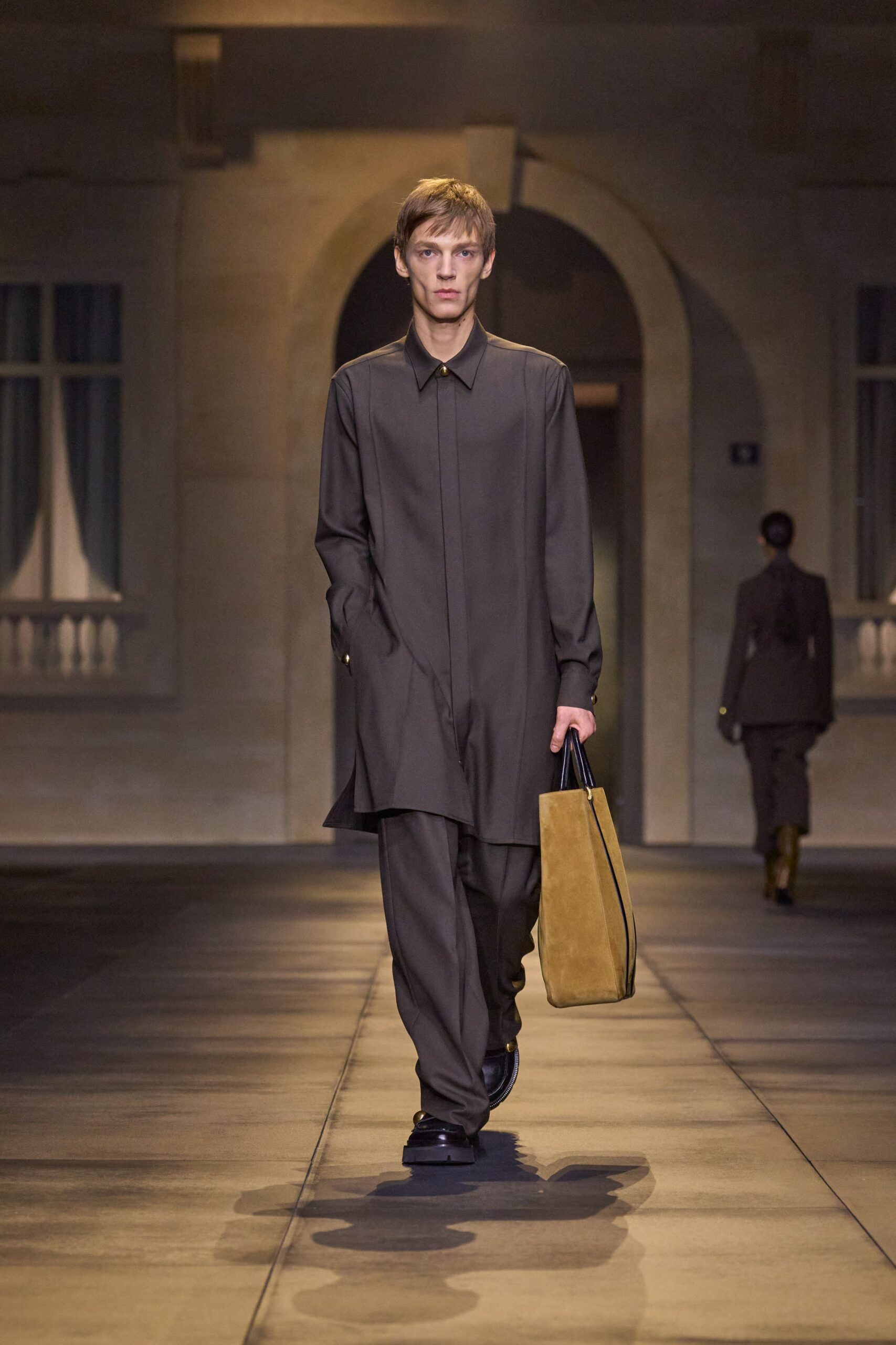 Long charcoal tunic shirt with a concealed placket and brass collar stud. Loose charcoal trousers breaking softly over the shoe. Tall camel suede tote with black rolled handles. Black leather shoes with a thick squared sole. Crossing a hushed hotel arcade before the shops unlock.