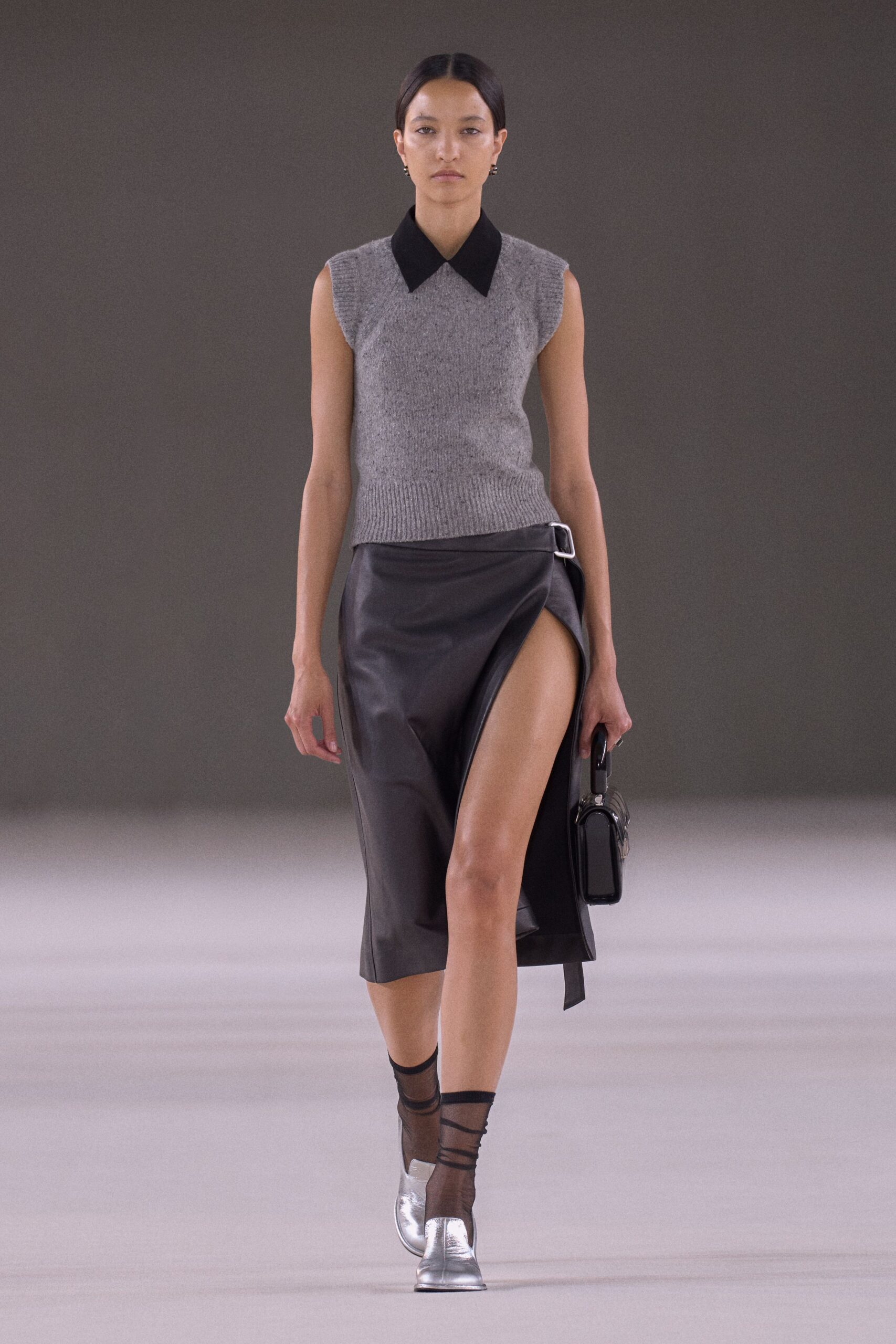Mottled grey knit sweater vest with a sharp black contrast collar. Black leather wrap skirt, thigh slit hooked into a silver side buckle. Glossy black box bag with a rigid top handle. Sheer black mesh, bands crossing the ankle. Silver leather flats with squared toes. For crossing a hotel lobby before anyone else is downstairs.