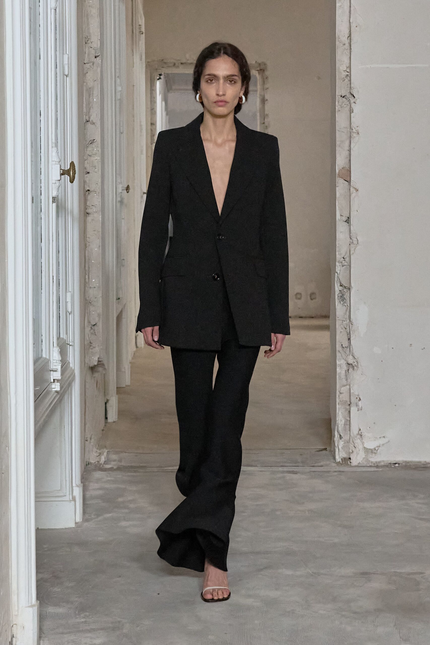 Long black tailoring with a plunging V and two glossy buttons. Black trousers breaking into a slight flare over the foot. Barely-there thong sandals with a slim pale strap. Small gold hoops. Whatever you wear back through an unfurnished apartment after signing the lease.