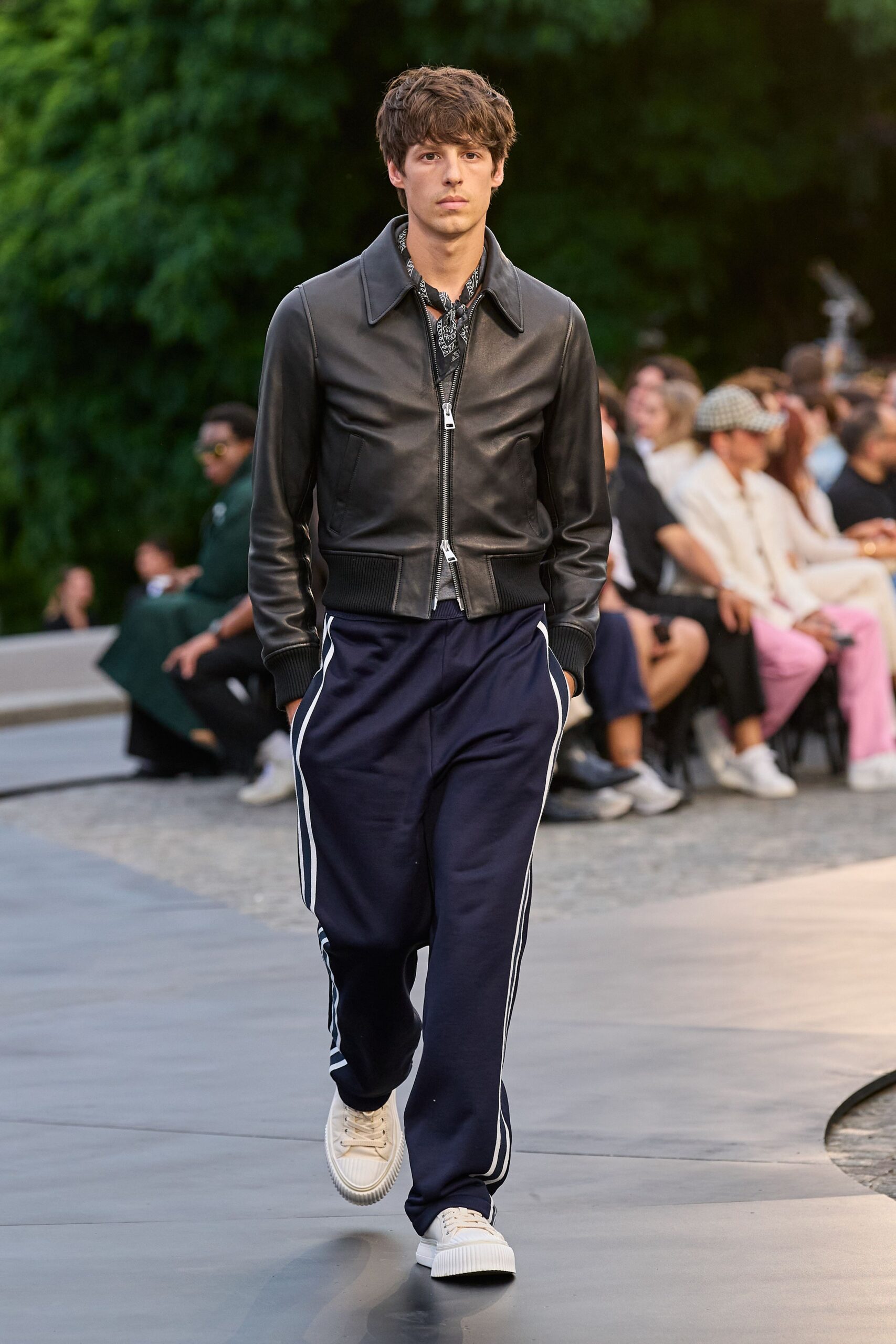 Black bandana scarf knotted close, white paisley peeking above the zip. Black leather blouson with a wide point collar and ribbed hem. Navy track pants with double white side stripes breaking at the cuff. Cream sneakers with ridged rubber foxing. Walking to dinner through a park while the heat is finally lifting.