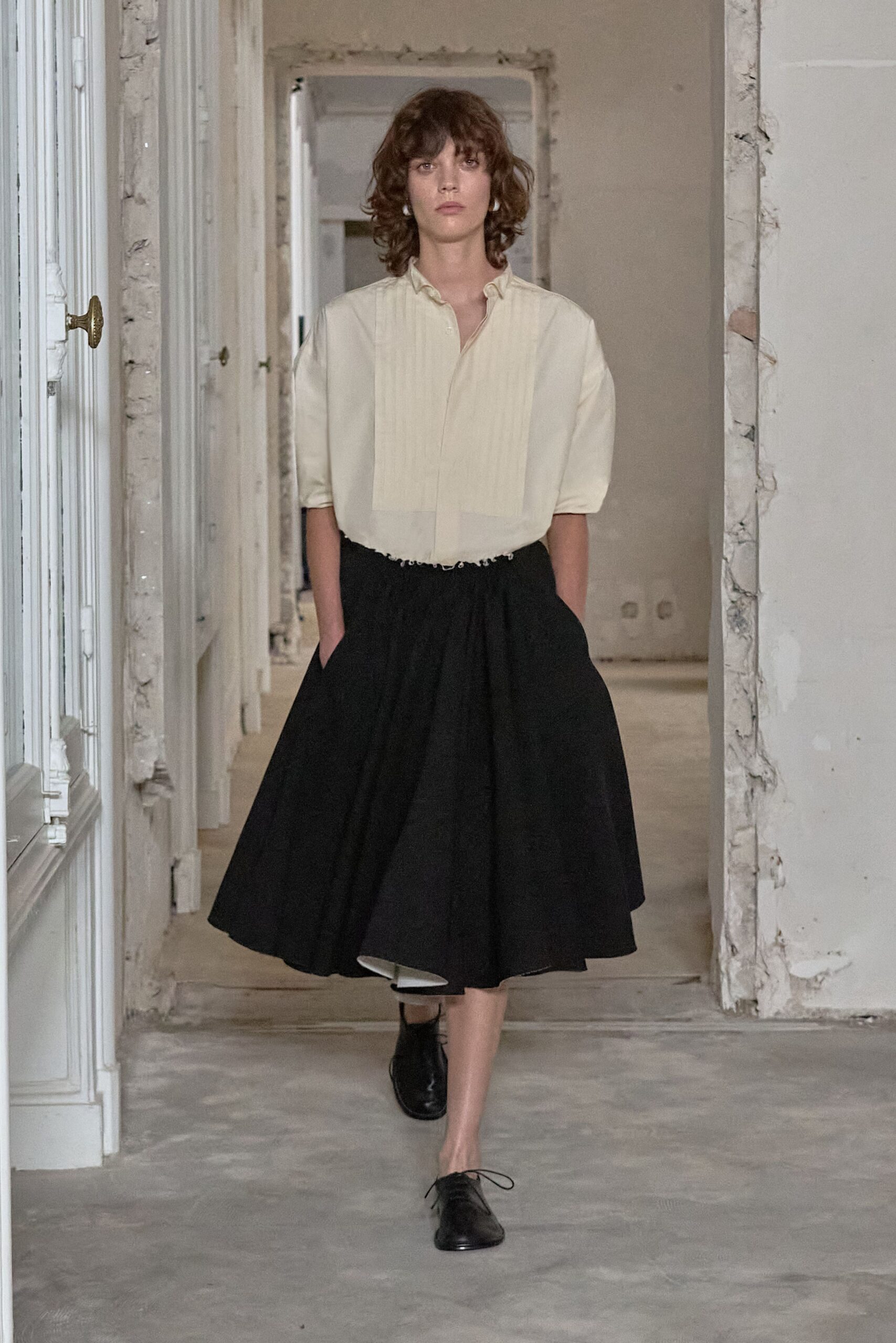 Cream blouse with a pintucked bib and frayed cropped hem. Black gathered midi skirt, wide cut swinging open to a pale lining. Black leather derbies with thin laces skimming the floor. Walking through an unfurnished apartment before the plaster dust settles.