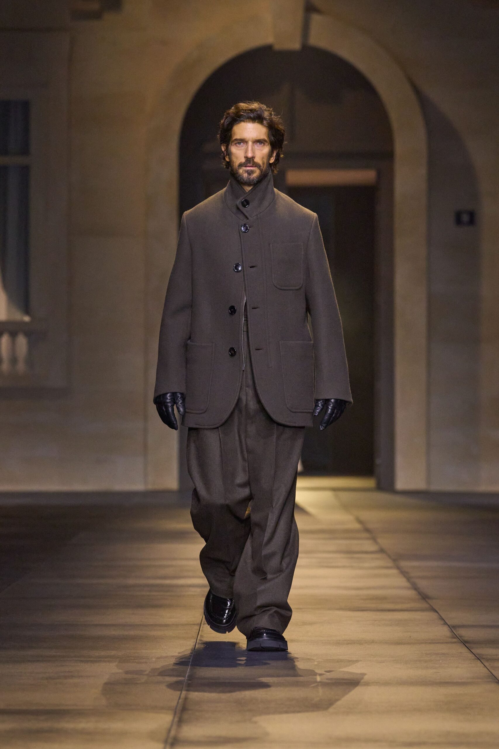 Taupe felted jacket with a funnel neck and rounded patch pockets. Loose brown wool trousers, front crease breaking softly over the shoe. Black leather gloves. Chunky black loafers with a blunt apron toe. Crossing a stone courtyard before the office radiators have clicked on.