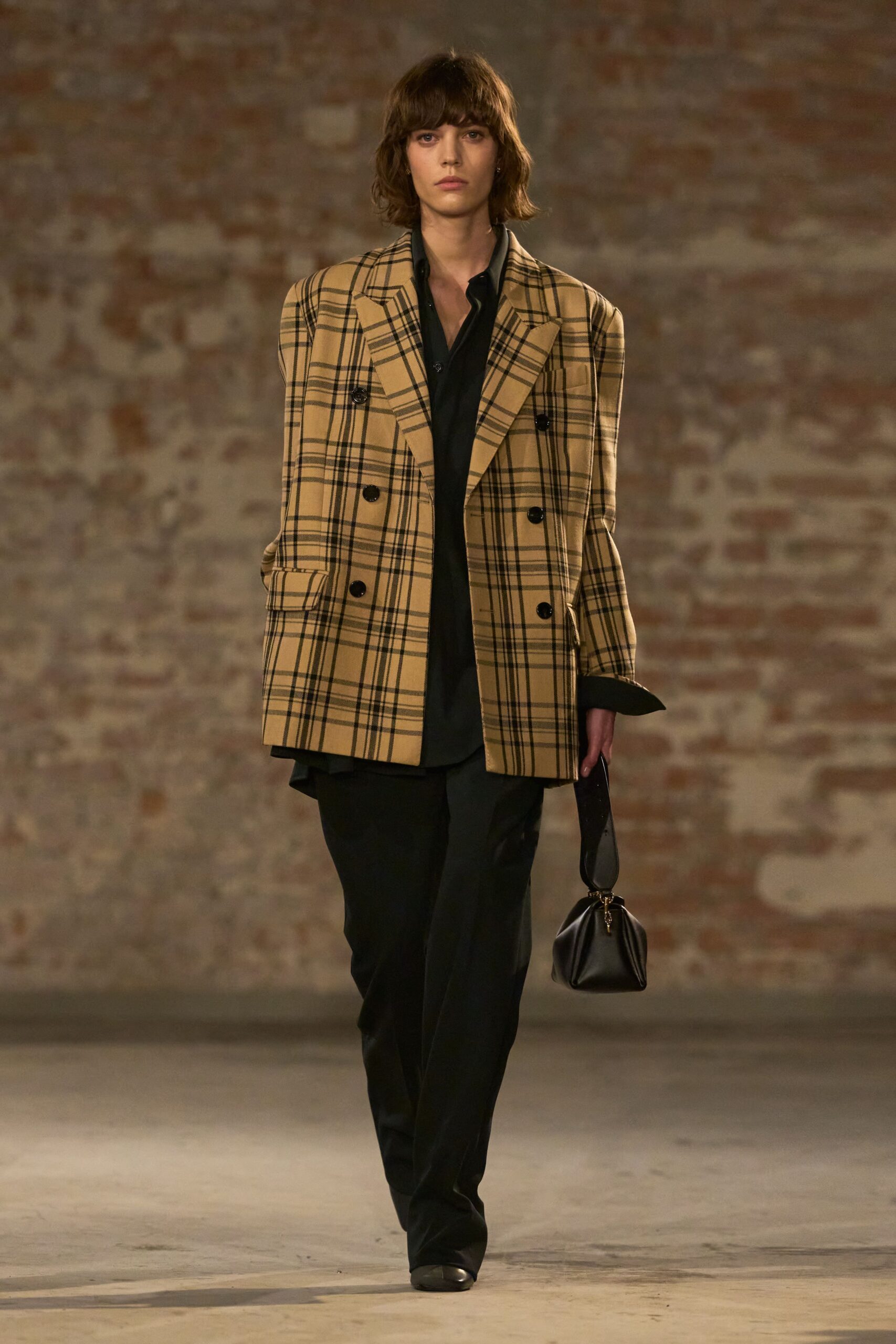 Camel plaid double-breasted blazer with squared shoulders and shiny black buttons. Black fluid shirt, open at the throat with long cuffs turned back. Black straight trousers breaking softly over the shoe. Small black pouch bag on a wide looped strap. Walking into a hotel bar before the rain lets up.