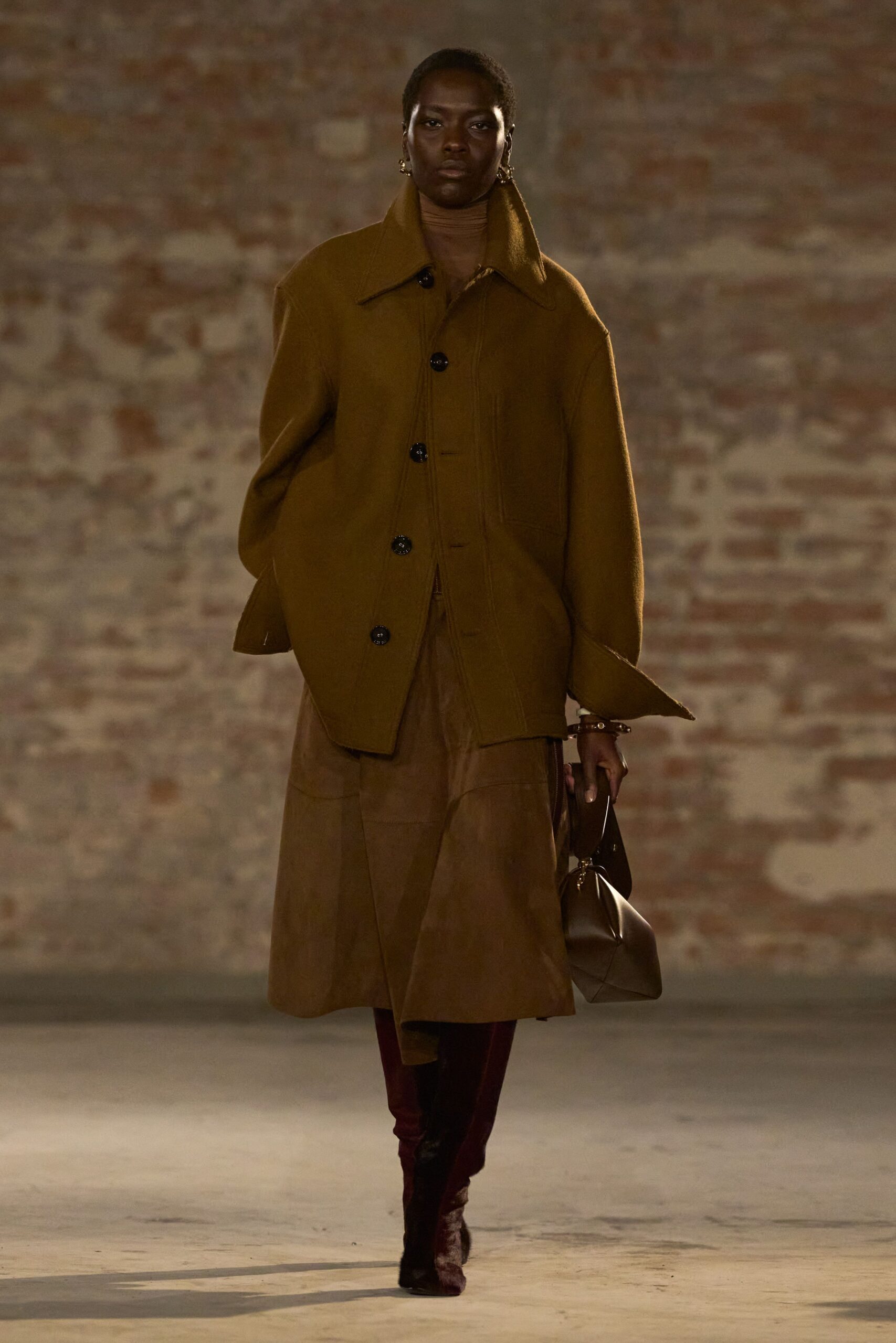 Oversized tobacco wool coat with a long collar and glossy black buttons. Fine brown knit turtleneck close at the throat. Cocoa suede midi skirt, paneled into a soft uneven flare. Dark leather pouch bag with a narrow wrist strap. Oxblood knee boots in crushed velvet. For crossing a cold courtyard with keys already in hand.
