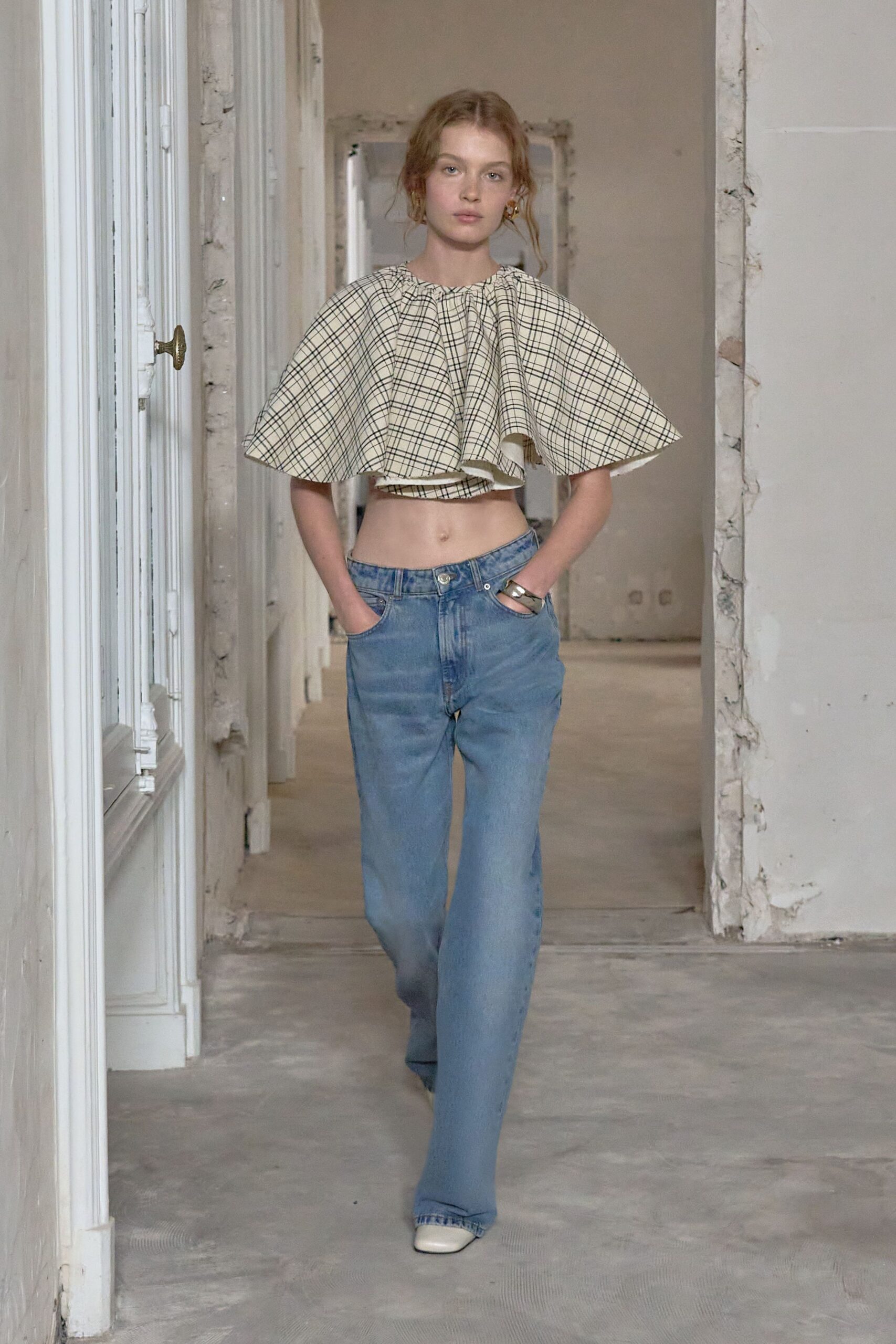 Cream cotton cape top in black windowpane checks, gathered tight at the neck. Faded blue jeans cut low on the hips, one pocket tugged open. Wide silver cuff at the wrist. Square-toe white boots peeking past the hem. Sunday apartment viewing, plaster dust still on the floorboards.