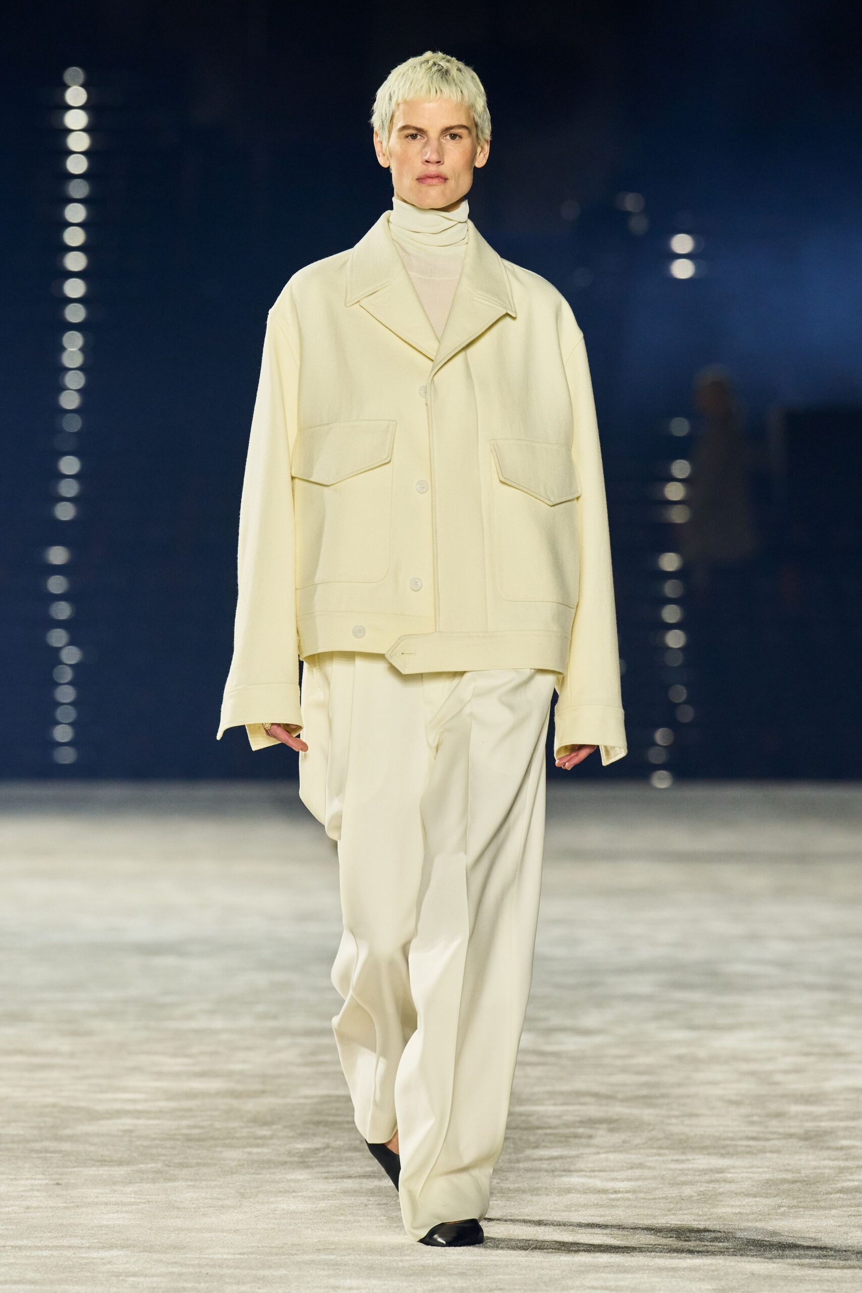 Cream fine-rib turtleneck stacked in soft folds at the throat. Oversized ivory wool jacket with wide collar and low flap pockets. Loose ivory trousers breaking over the shoe in heavy drape. Black pointed boots peeking out beneath the pooled hem. Early ferry crossing, deck lights still on against the water.