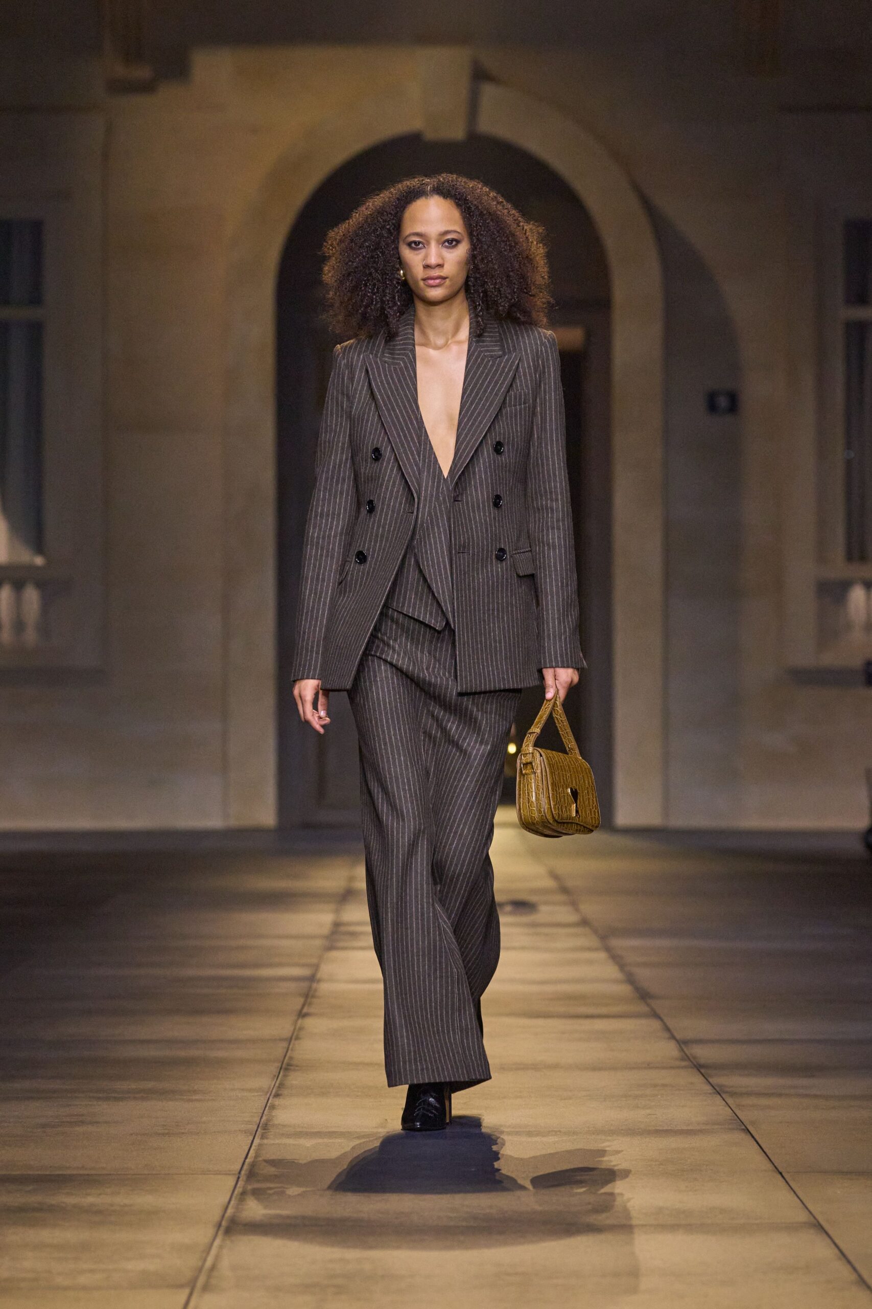 Brown pinstripe blazer cut double-breasted, worn bare with a plunging lapel. Matching wide-leg trousers breaking softly over the shoe. Ochre crocodile-embossed shoulder bag with a cutout clasp. Black square-toe shoes. Crossing a stone courtyard just before the dinner reservation turns useful.