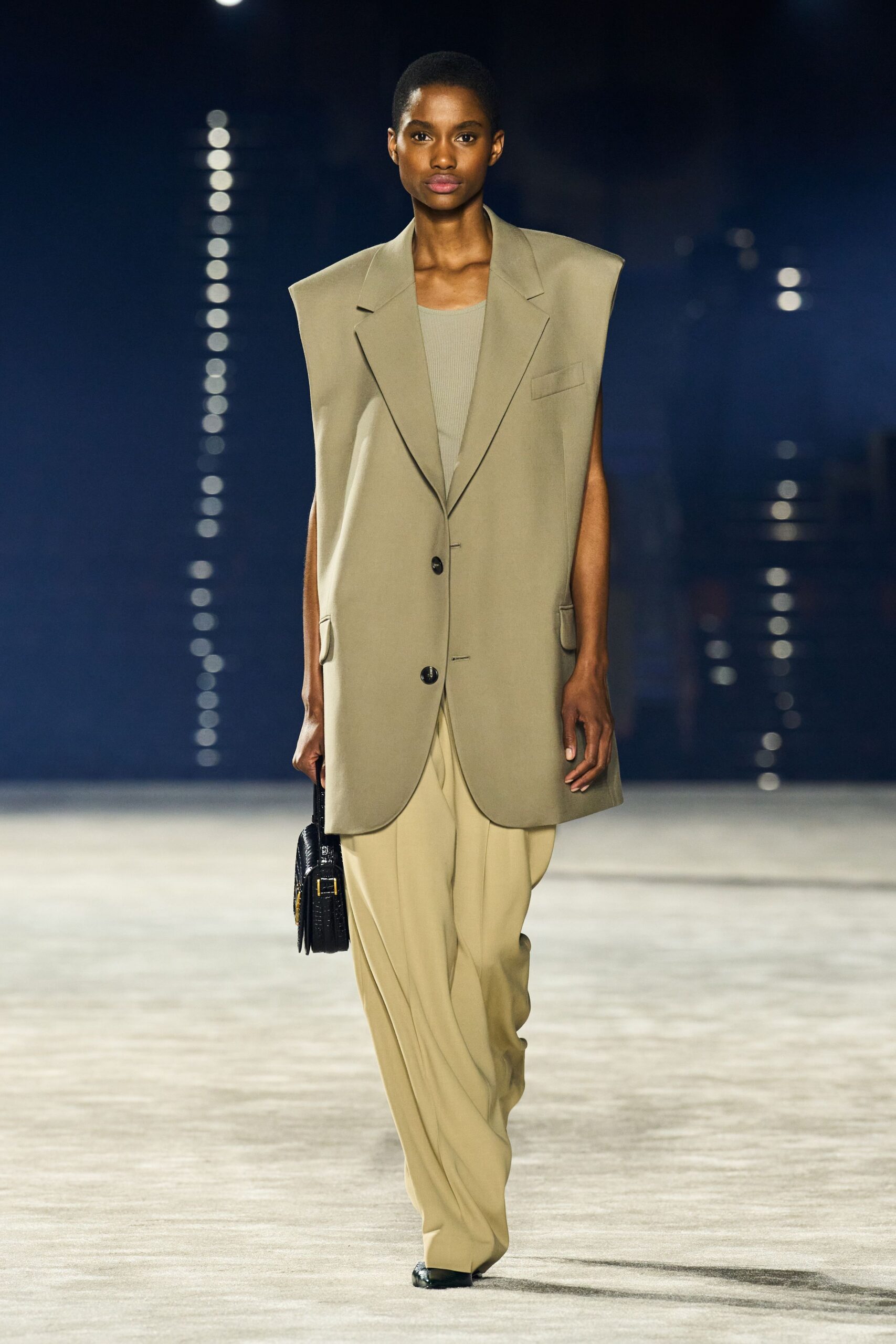 Oversized putty suiting vest with squared shoulders and two dark buttons. Fine ribbed tank in pale sage. Buttercream wide-leg trousers breaking into soft folds at the shoe. Black croc-embossed portfolio with a gold buckle edge. Black pointed shoes. For crossing a near-empty airport before the coffee carts open.