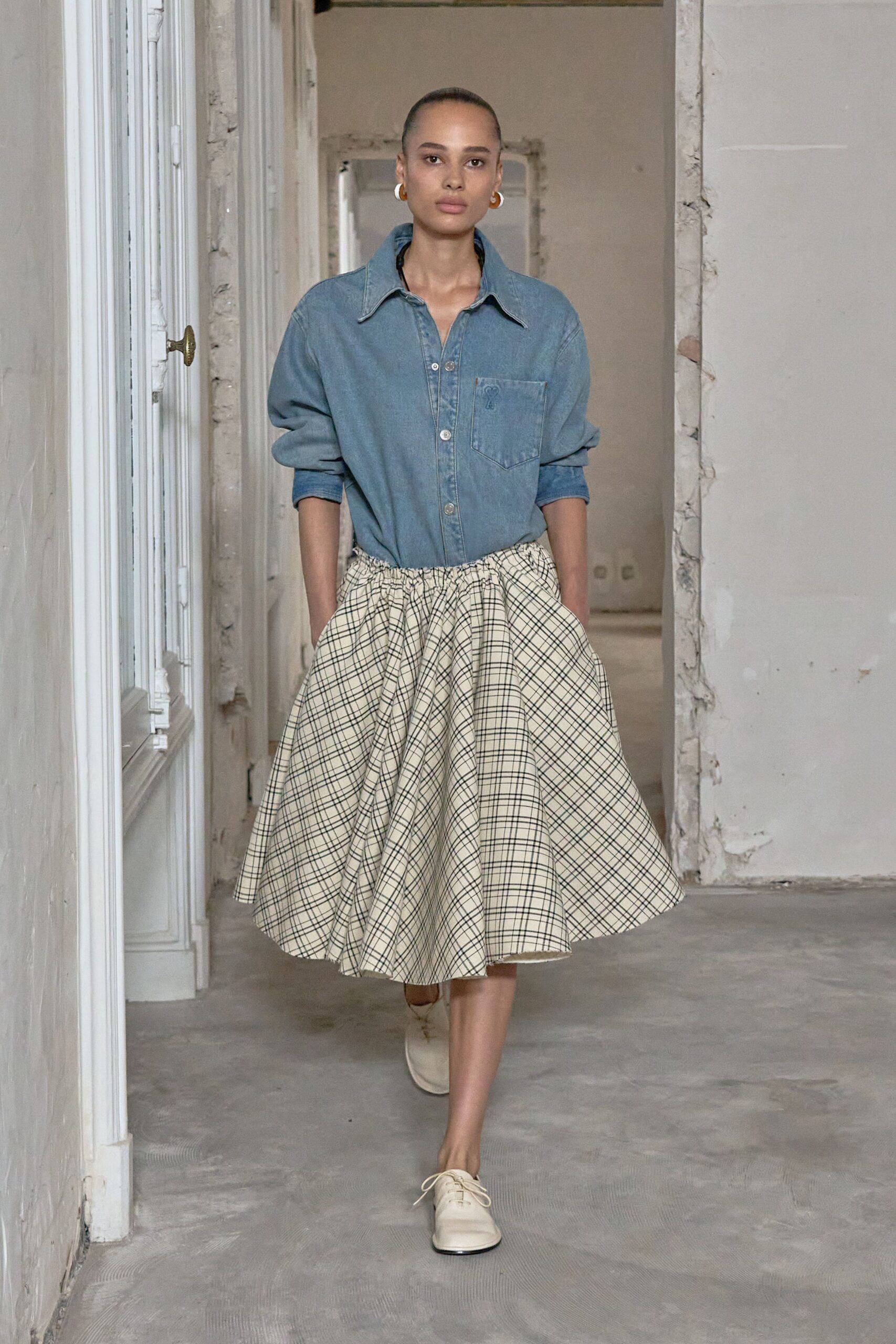 Small amber resin hoops edged in gold. Washed blue chambray shirt, broad collar and a single chest pocket. Cream full skirt in black windowpane cotton, gathered hard at the waist. Bone leather derbies with thin round laces. Walking through a half-renovated apartment before the curtains go up.