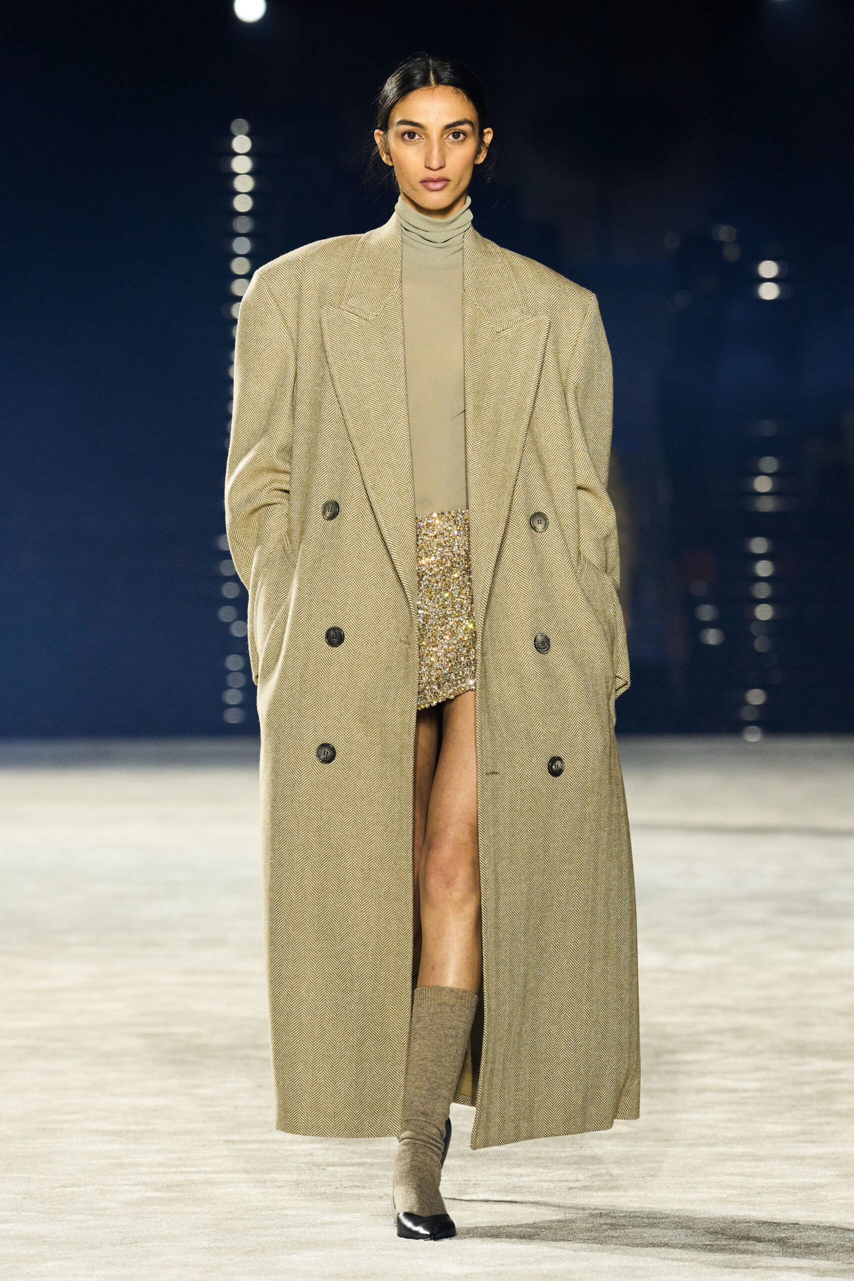 Oversized taupe herringbone coat, dropped shoulders and dark double-breasted buttons. Fine ribbed beige turtleneck rolled softly at the throat. Gold paillette mini flashing beneath the open coat. Taupe knit sock boots with black dipped square toes. Walking out for cigarettes after midnight in a hotel that still has a pianist.