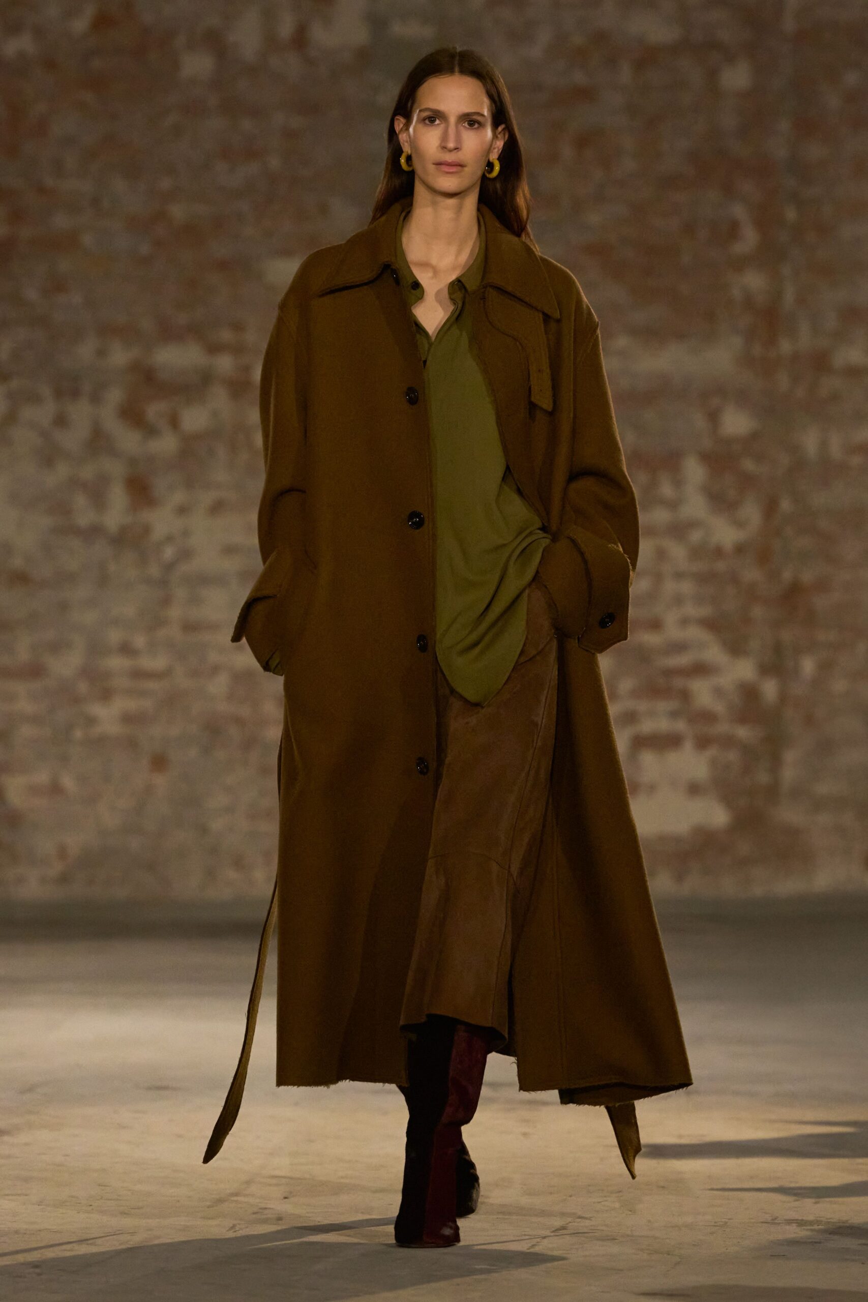 Chunky chartreuse hoops. Floor-length walnut wool coat, raw hem skimming the concrete. Olive blouse left loose, placket open at the throat. Chestnut leather midi skirt cut in curved panel seams. Oxblood knee boots, square toes darkened at the vamp. Walking into a draughty town hall before the auction starts.