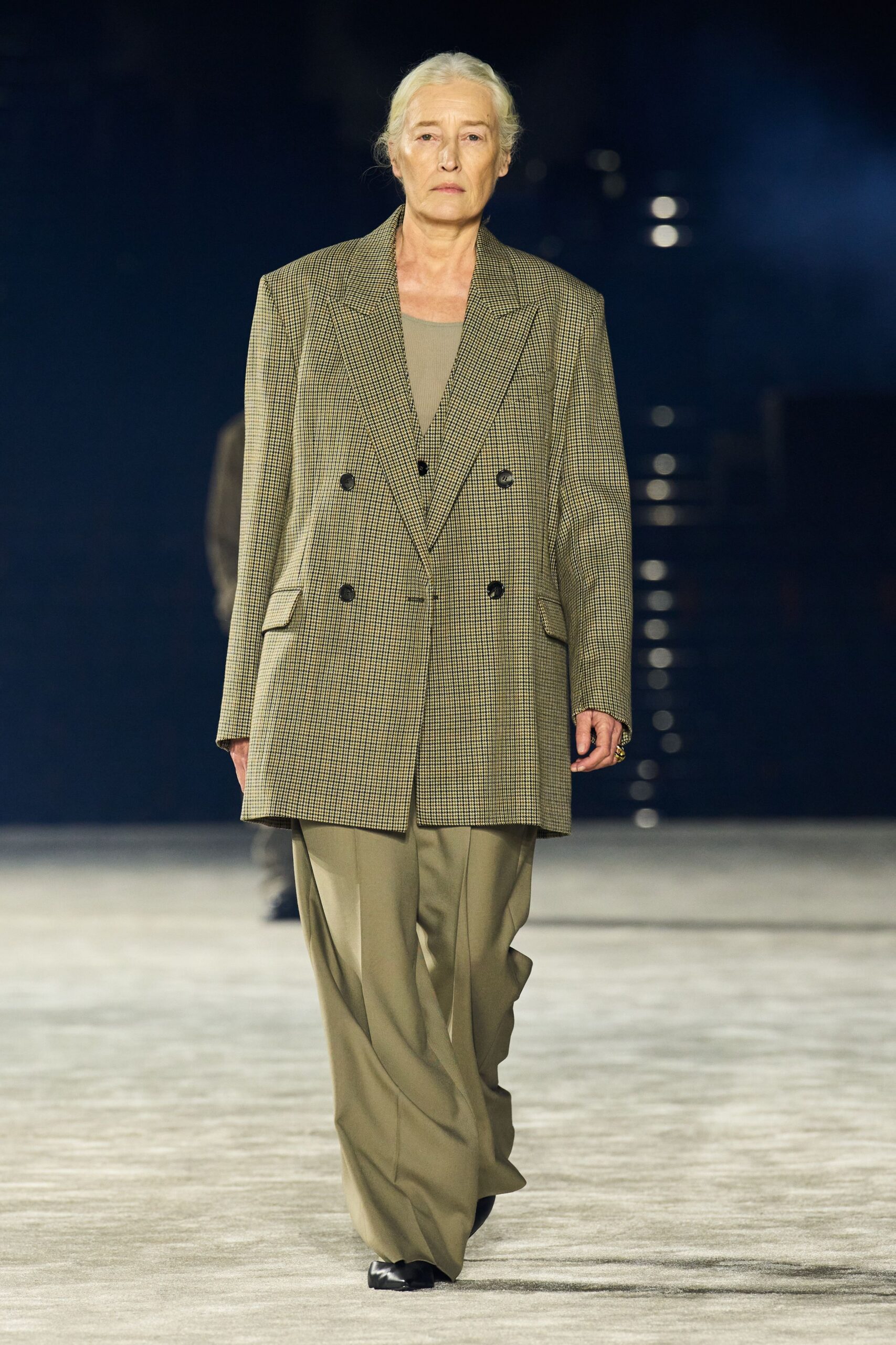 Long double-breasted blazer in tiny taupe check, shoulders cut broad. Taupe crewneck layer, plain and close to the body. Wide khaki trousers with a pressed front crease breaking over the shoe. Black square-toe shoes. For crossing the hotel lobby before dawn with yesterday's keycard still working.