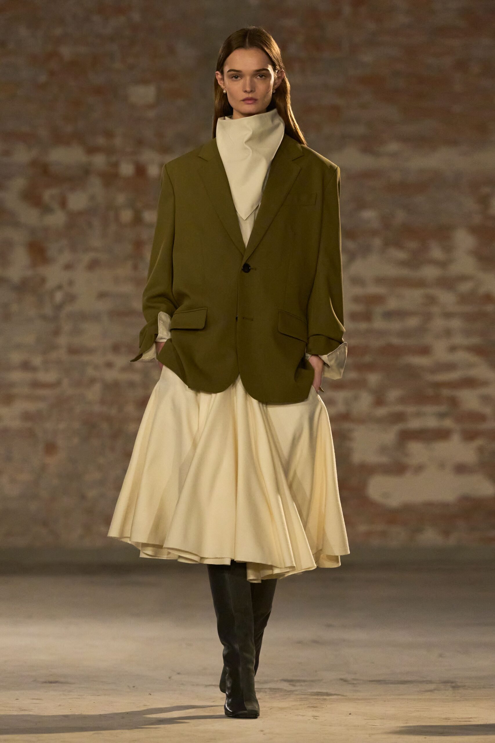 Olive wool blazer cut loose, with low flap pockets and a single dark button. Ivory dress with a sculptural funnel neck and soft handkerchief hem. Black knee-high leather boots, narrow and plain under the skirt. Crossing a museum courtyard just before the rain starts.