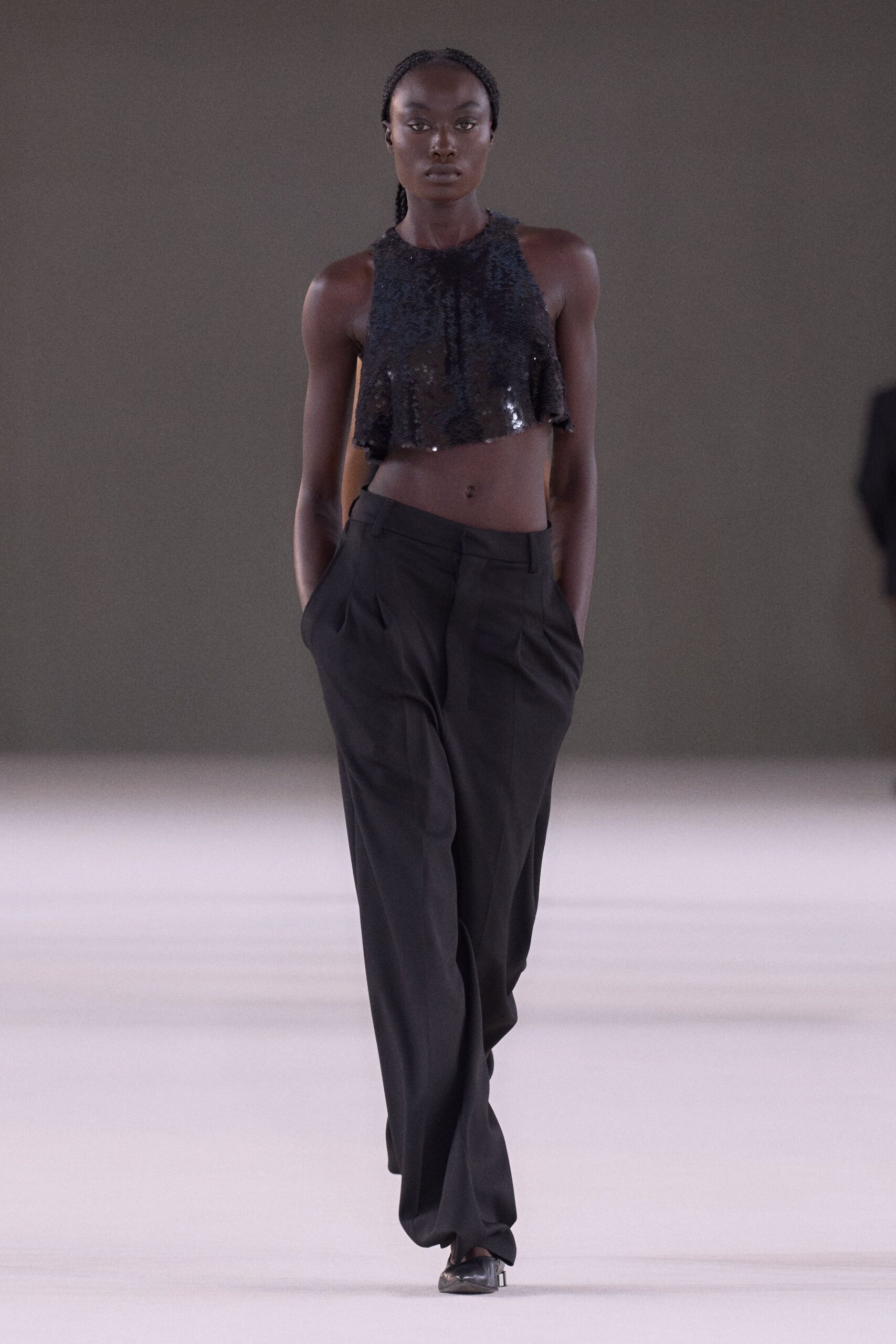 Black sequin halter crop top, raw fringe flickering along the hem. Low-slung black wool trousers with deep pleats and hands-buried pockets. Pointed black pumps peeking from the pooling break. After midnight cleanup, confetti still stuck to the dance floor.
