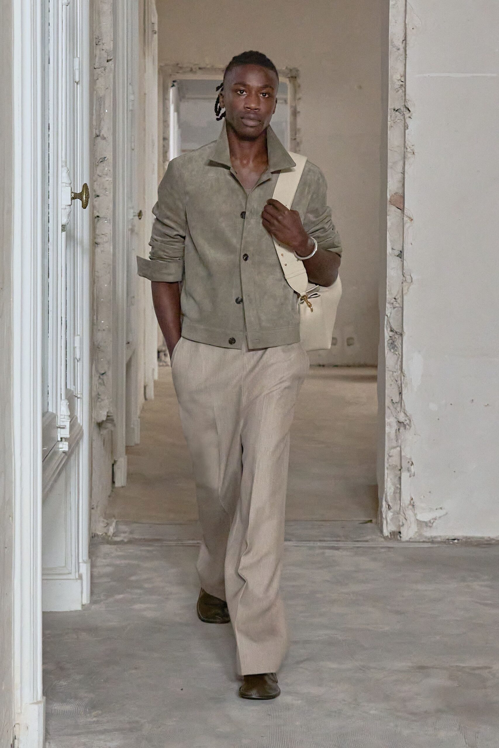 Moss suede jacket with a wide collar and sleeves pushed up. Stone straight-leg trousers with a soft, dry drape. Cream leather shoulder bag on a broad strap with brass clip. Plain silver bangle. Dark square-toe shoes. Walking through a half-renovated apartment before the paint smell lifts.