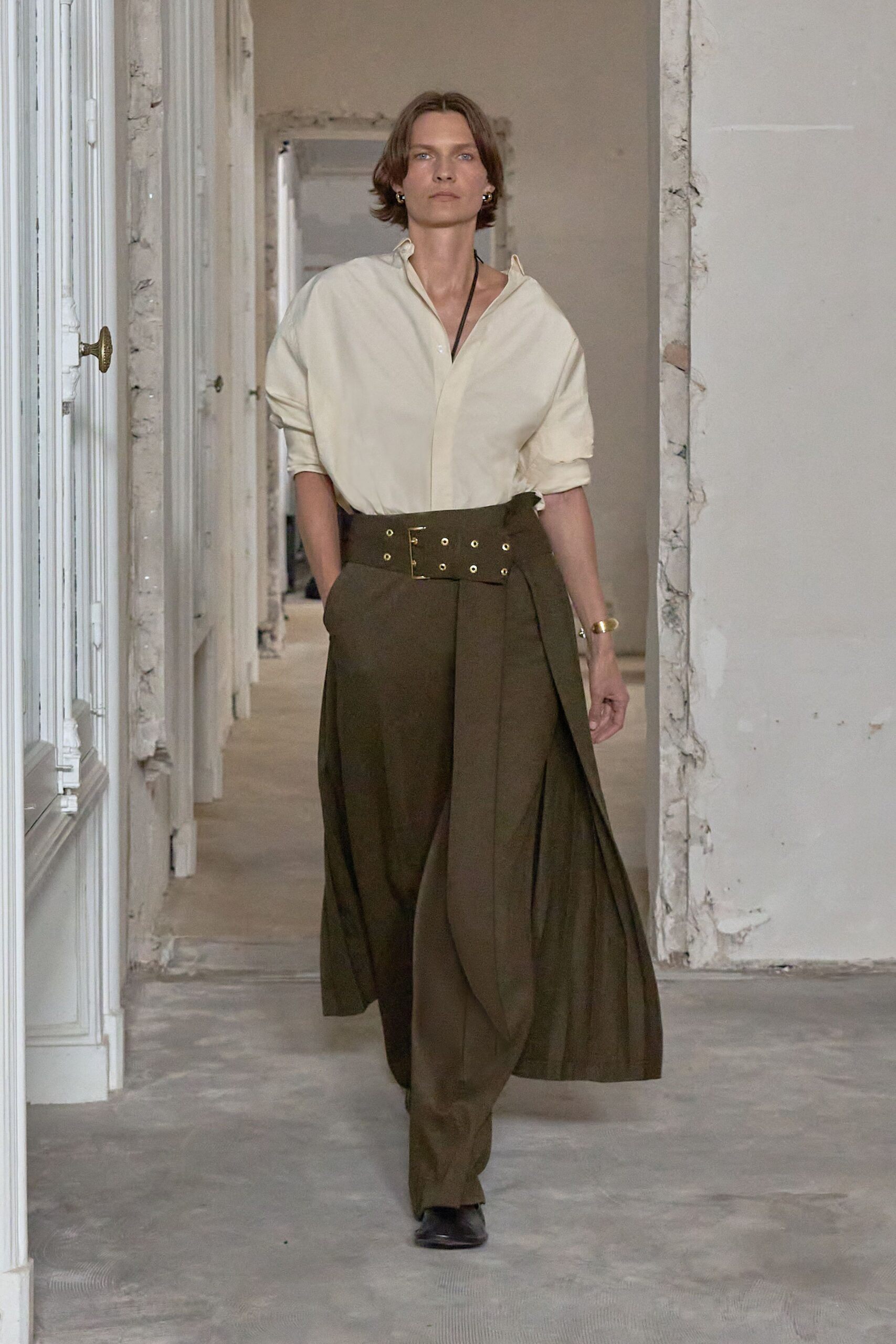 Ivory cotton poplin shirt, collar sprung open and sleeves loosely rolled. Dark olive wide-leg trousers hidden under an apron-like wrap panel. Broad self belt punched with twin rows of brass eyelets. Black square-toe boots peeking under the hem. For crossing a half-renovated apartment before the movers arrive.