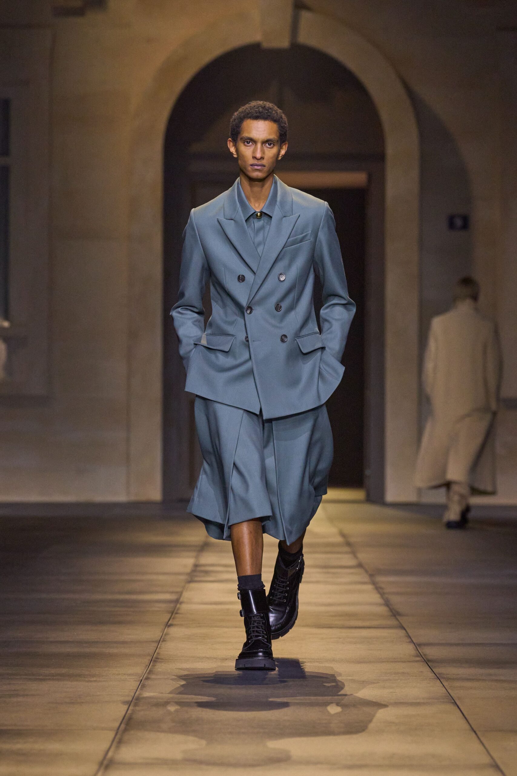Dusty blue double-breasted blazer, one pocket flap kicked outward. Matching shirt fastened high with a single brass dome. Knife-pleated culottes breaking into soft panels below the knee. Black ribbed socks. Black leather combat boots with a broad ankle strap. Crossing a stone cloister just before the bell.
