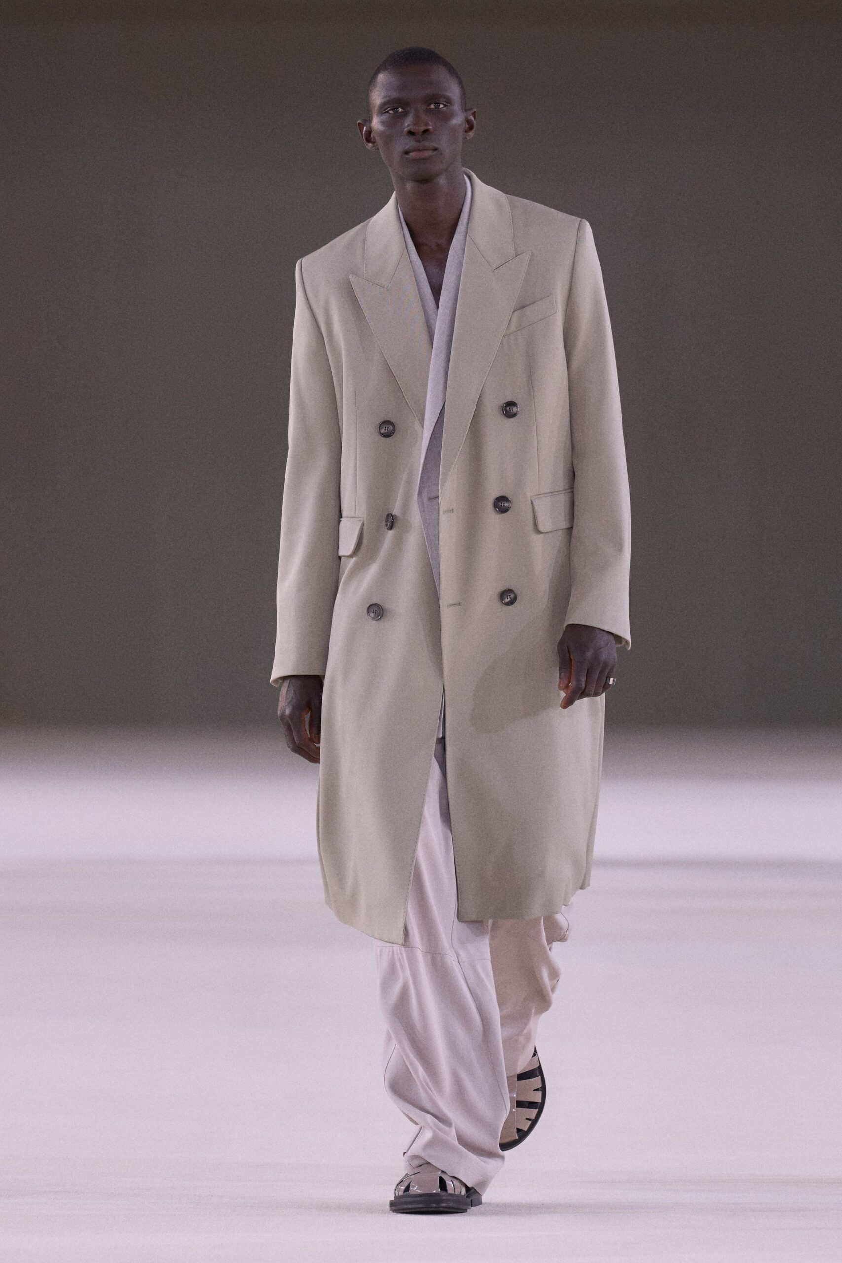 Long oyster double-breasted coat with peaked lapels and smoky horn buttons. Pale draped V-neck layer disappearing deep beneath the coat. Chalk satin trousers breaking softly over the foot. Black-and-cream cutout sandals with squared soles. Crossing a silent hotel lobby before anyone else is awake.