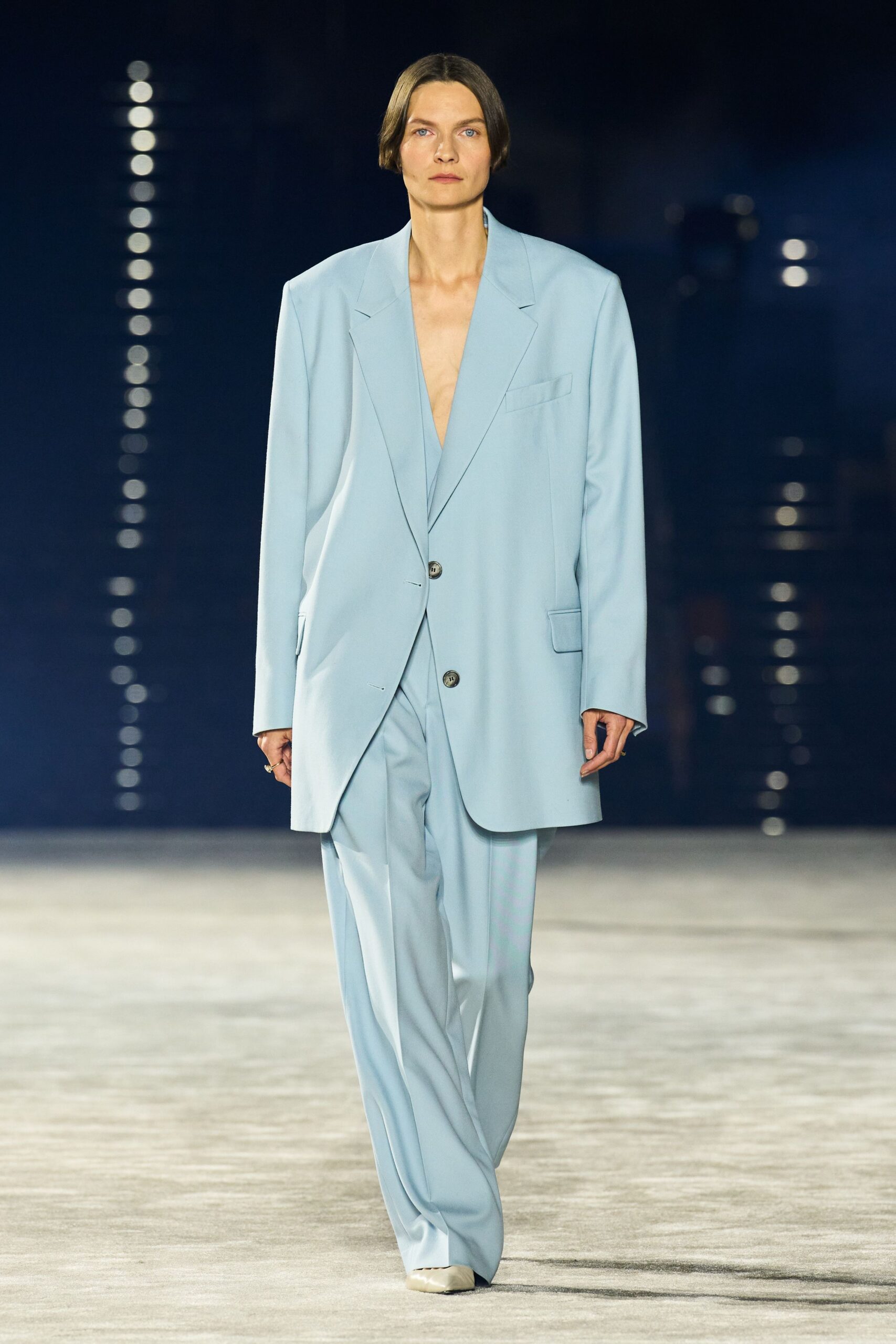 Powder-blue blazer cut oversized, with peaked lapels and low mottled buttons. Matching plunging inner layer, almost disappearing into the jacket opening. Loose powder-blue trousers breaking into soft folds over square-toe shoes. Cream square-toe shoes. Early check-in at a glassy airport hotel, before the lobby fills.