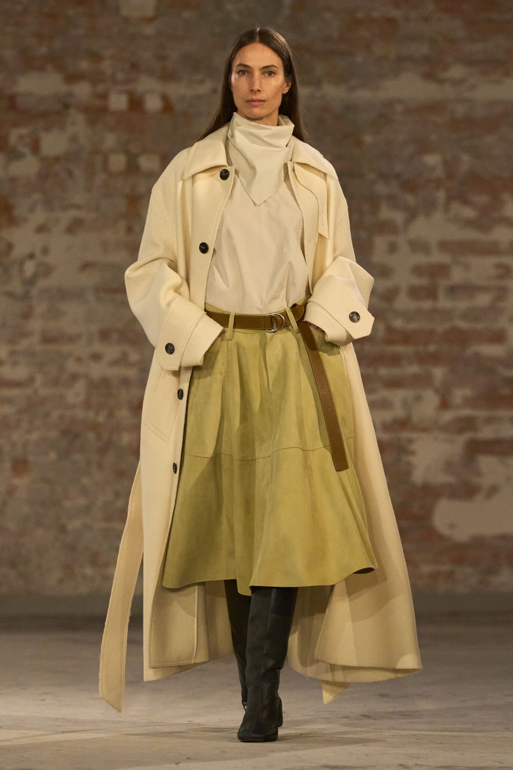 Cream double-face coat with oversized cuffs and black horn buttons. Ivory high-neck top, folded into a soft scarf-like bib. Olive paper-cotton skirt, lightly gathered under a dropped seam. Brown leather belt with a silver D-ring and long tail. Black knee boots. For crossing a museum courtyard in wind that keeps lifting the hem.