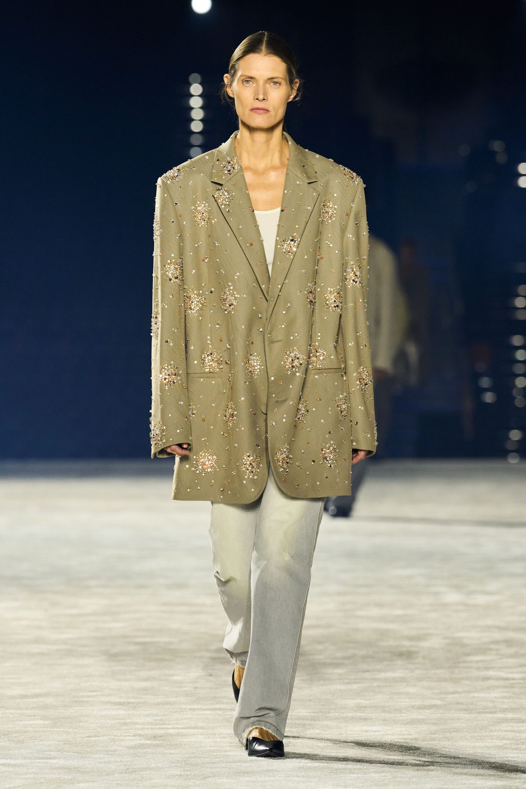 Oversized khaki blazer scattered with clustered crystal bursts and long sleeves. Ivory scoop-neck tank in fine rib knit. Pale stone wide-leg trousers breaking softly over the shoe. Black pointed pumps with a flash of gold at the vamp. Hotel ballroom after midnight, confetti ground into the carpet.