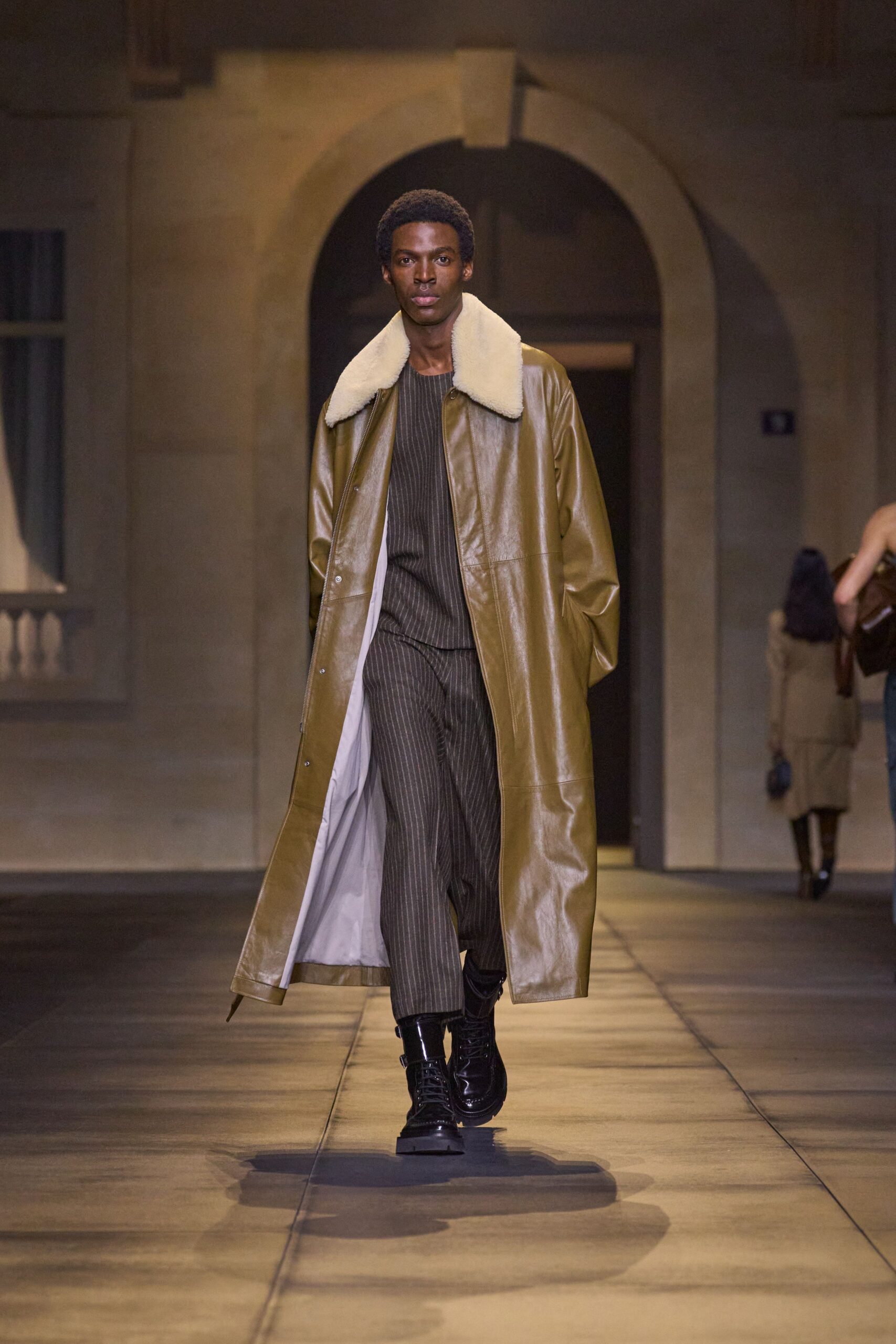 Olive leather coat with an oversized cream shearling collar. Dark ribbed knit top, cut plain beneath the heavy outer layer. Charcoal pinstriped trousers falling straight to the ankle. Black patent lace-up boots with a strap buckled at the shaft. Crossing a stone cloister just before the building opens to visitors.