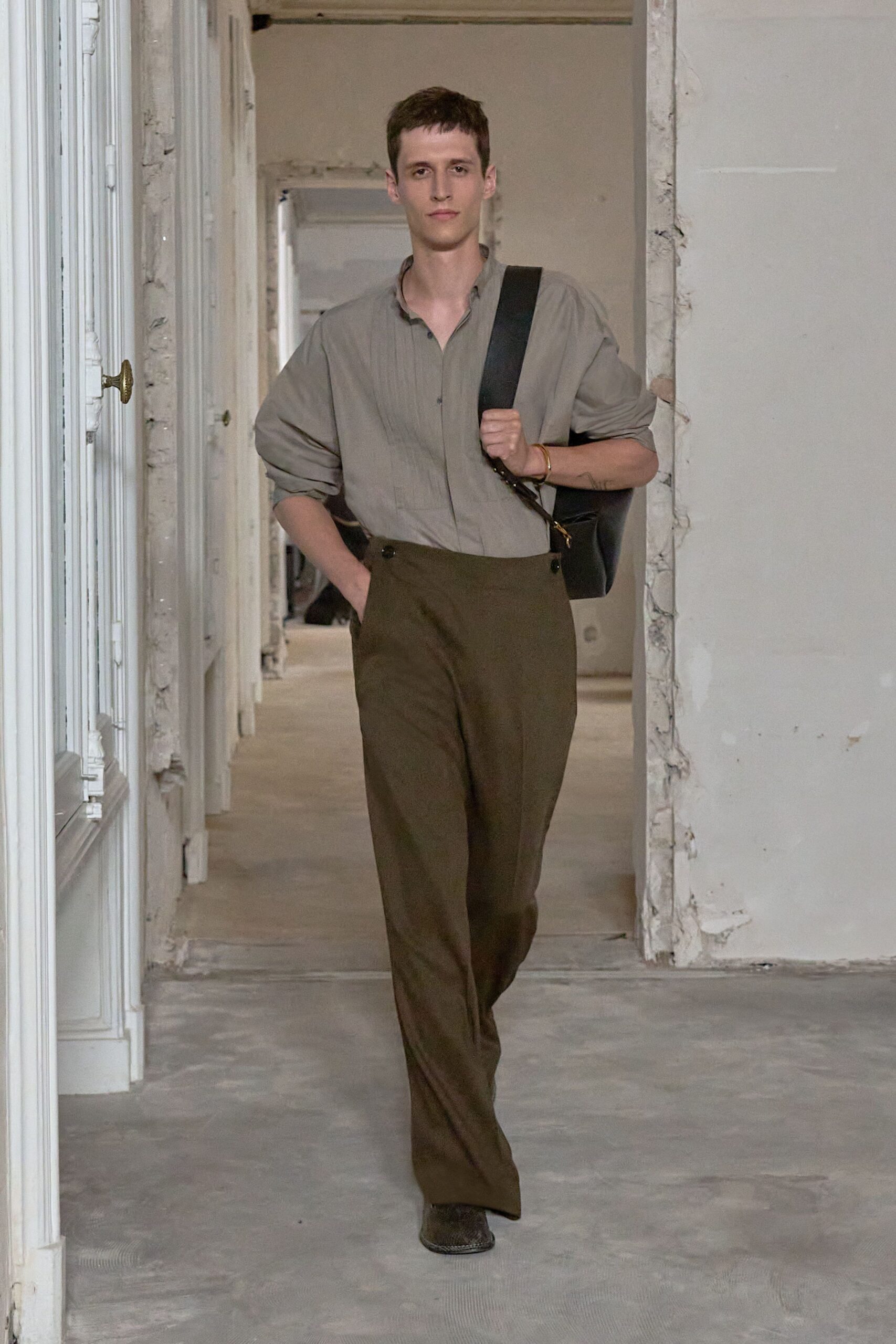 Stone-grey shirt with a soft band collar and narrow pintucks. Black leather shoulder bag on a broad strap, tucked underarm. Thin brass bangle. Olive high-waisted trousers with side buttons at the waistband. Dark snip-toe boots peeking from the hem. Walking through an empty apartment before the paint smell lifts.