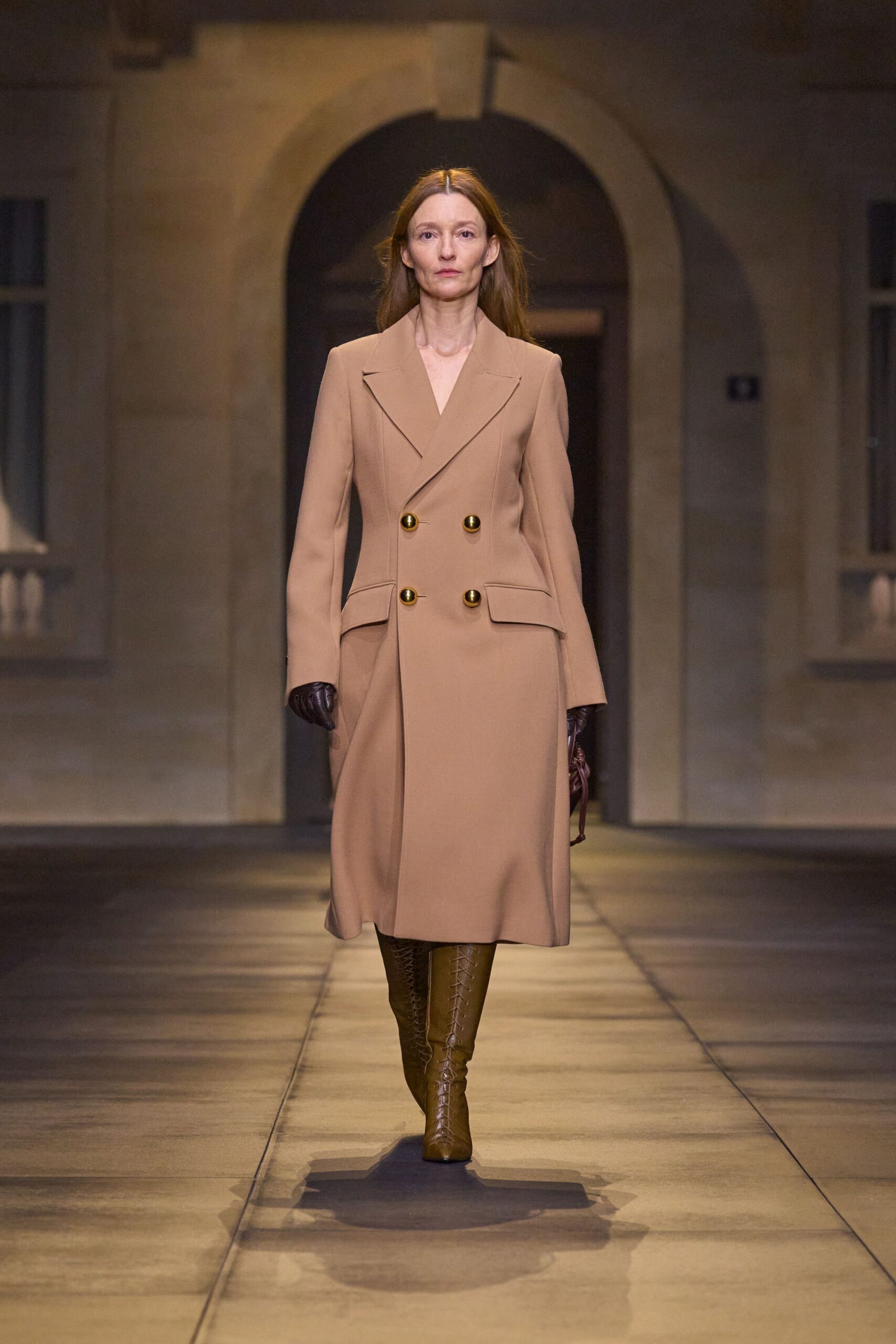 Camel double-breasted coatdress, four burnished dome buttons and slanted flap pockets. Dark oxblood leather gloves. Chestnut leather knee boots, ladder lacing running straight up the front. Crossing a stone courtyard before the hearing starts.