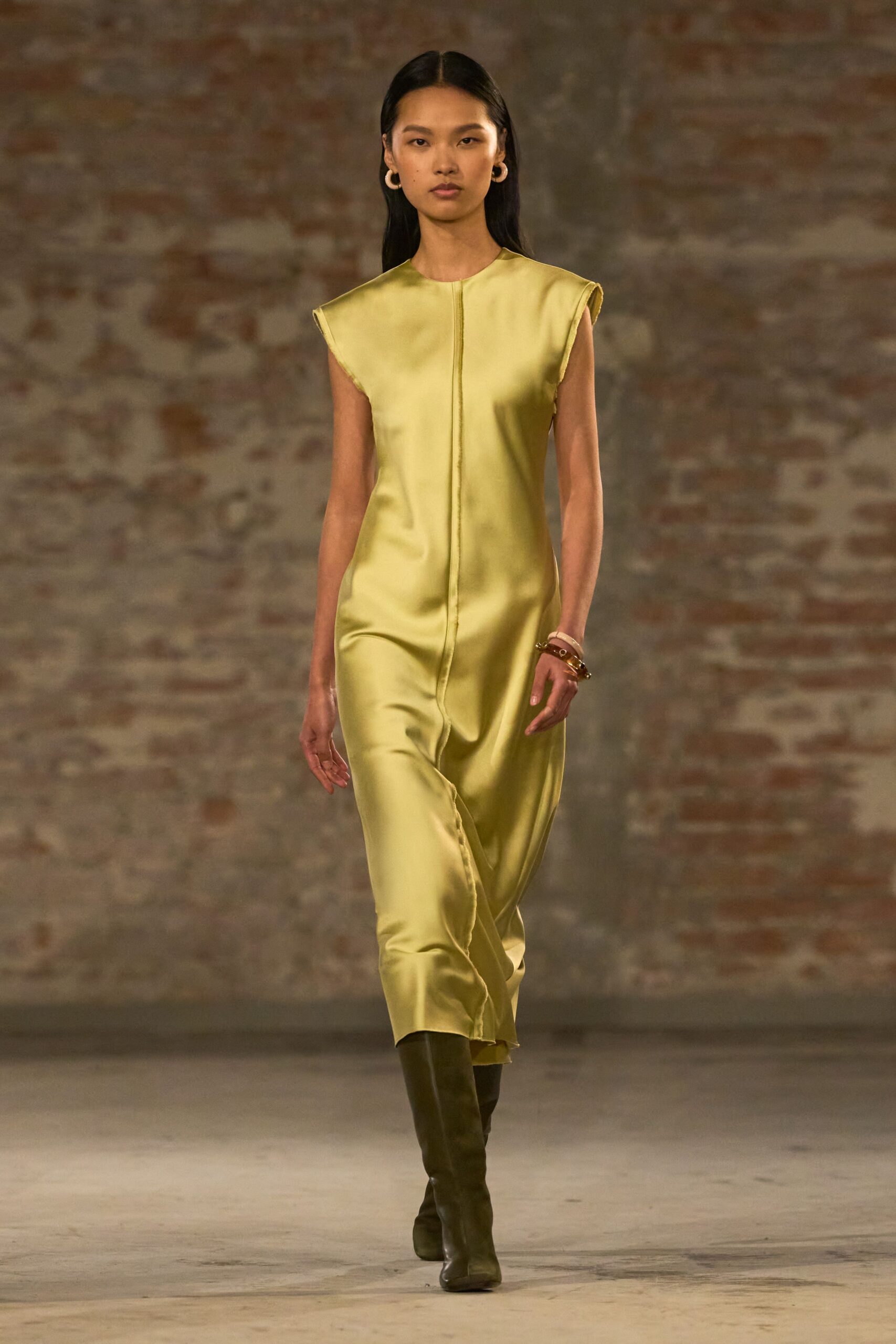 Chartreuse satin column dress with a raised center seam and raw cap edges. Stacked resin bangles in ivory and amber. Chunky pale hoops hugging the lobe. Olive leather knee boots with squared toes. Walking across a museum courtyard just after the rain lifts.
