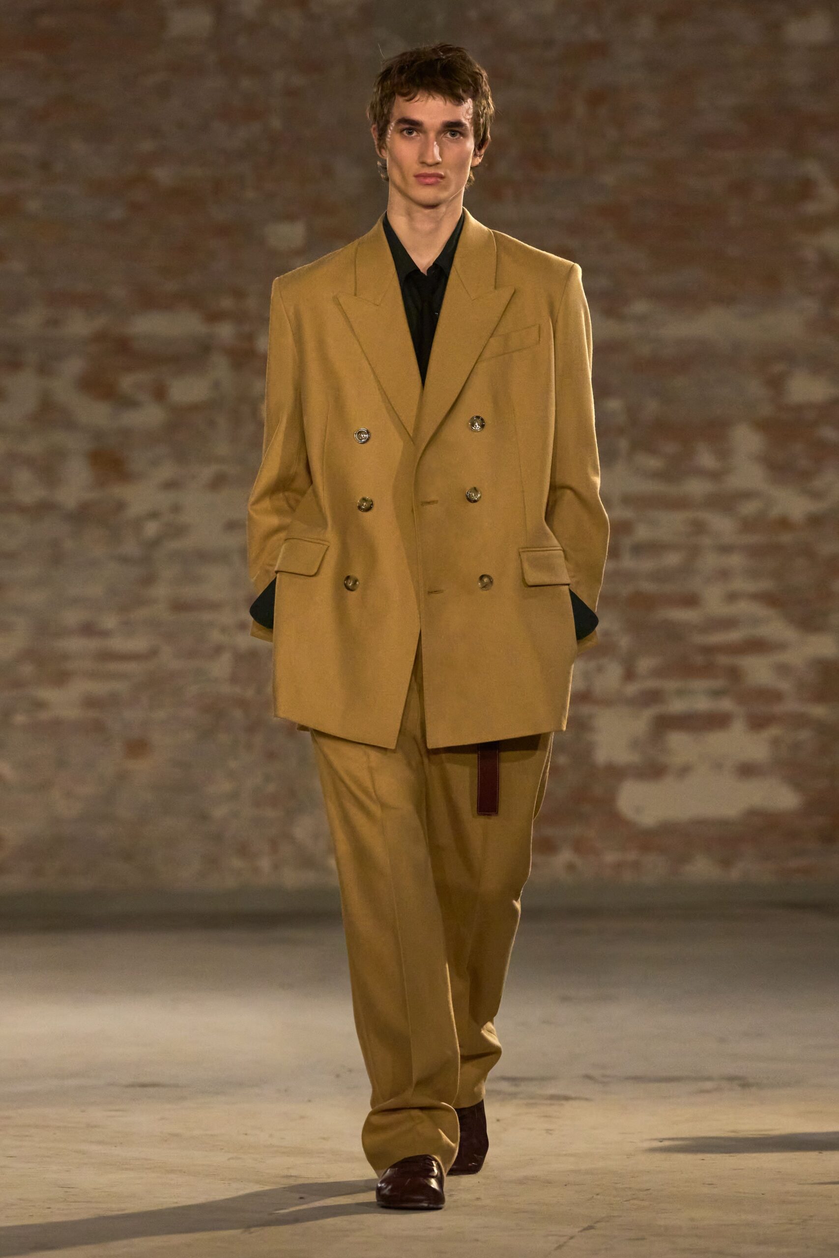 Camel double-breasted blazer with sharp peak lapels and marbled gold buttons. Black shirt with a soft open collar and cuffs hanging long. Matching camel trousers falling straight, a burgundy strap peeking at the fly. Dark oxblood leather shoes with squared toes. For crossing a drafty hall after the deal is finally signed.