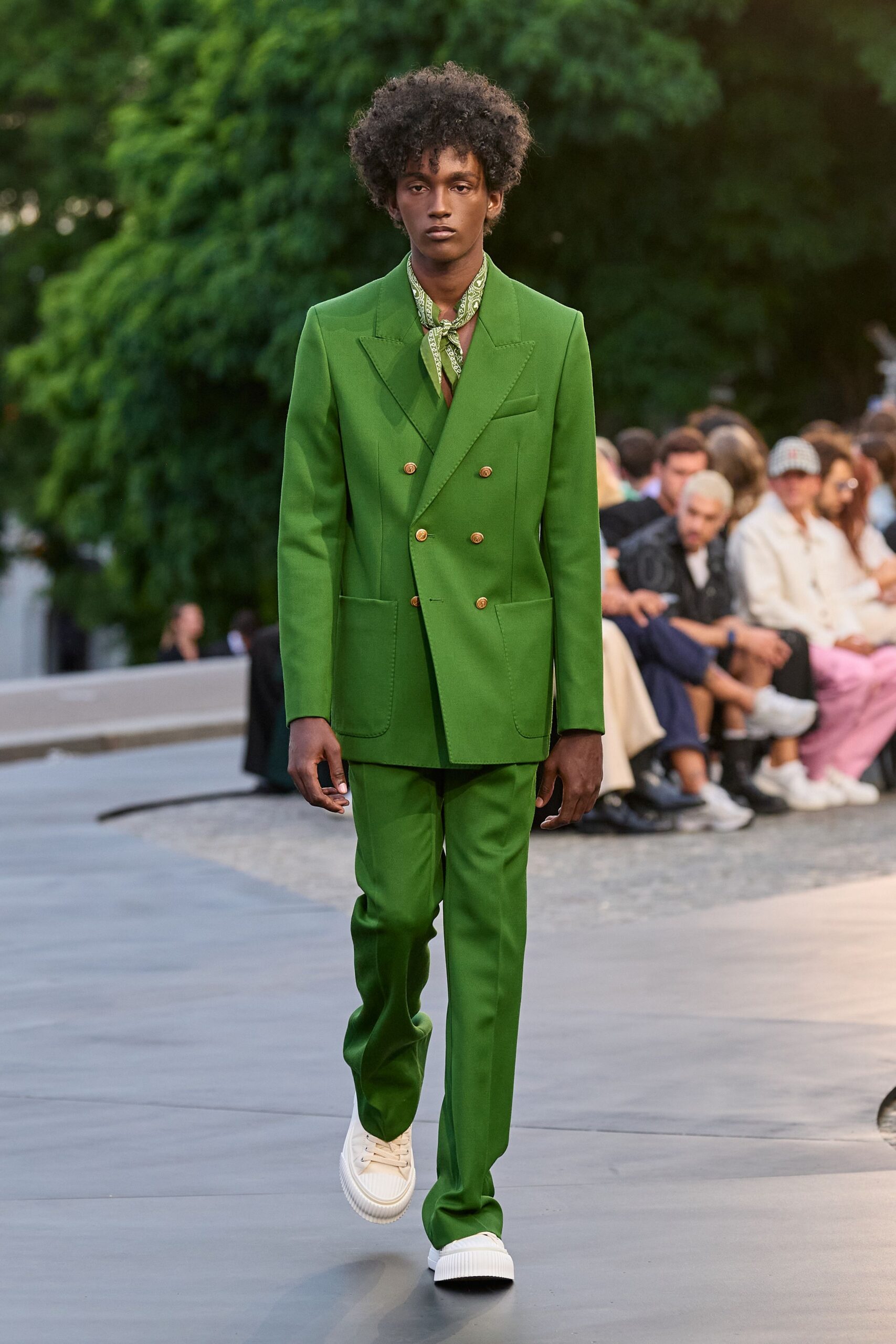 Pale sage paisley scarf knotted close at the throat. Grass-green tailoring with peaked lapels, gold crested buttons and patch pockets. Matching straight trousers breaking softly over the shoe. Cream platform sneakers with ribbed foxing. Crossing a formal garden just after the sprinkler cycle ends.
