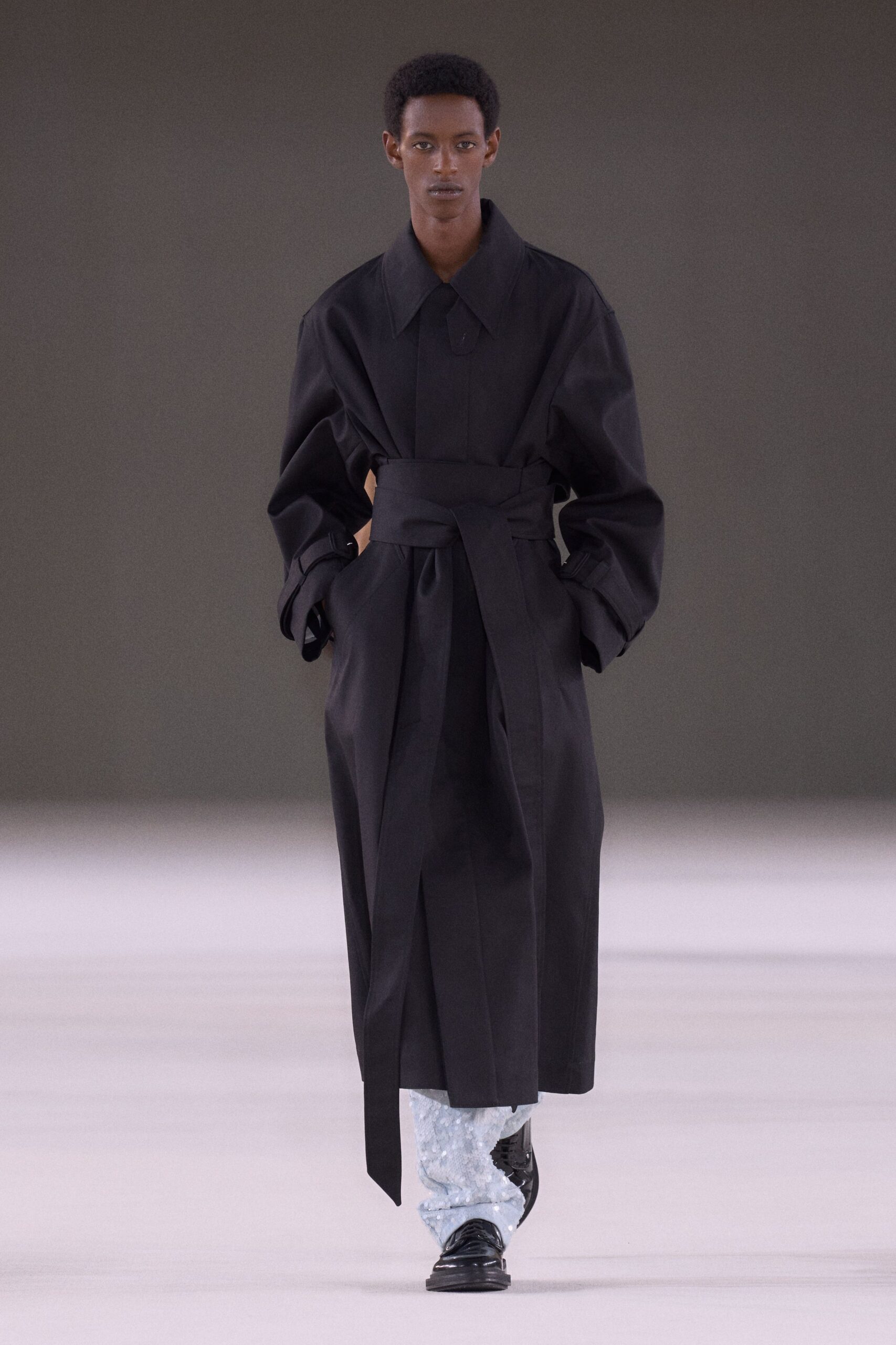 Black cotton coat with a huge collar and obi-wrap waist. Icy sequined trousers bunched over the shin in wet-looking clusters. Black patent loafers with thick squared soles. Crossing a hotel lobby at dawn with yesterday still on.