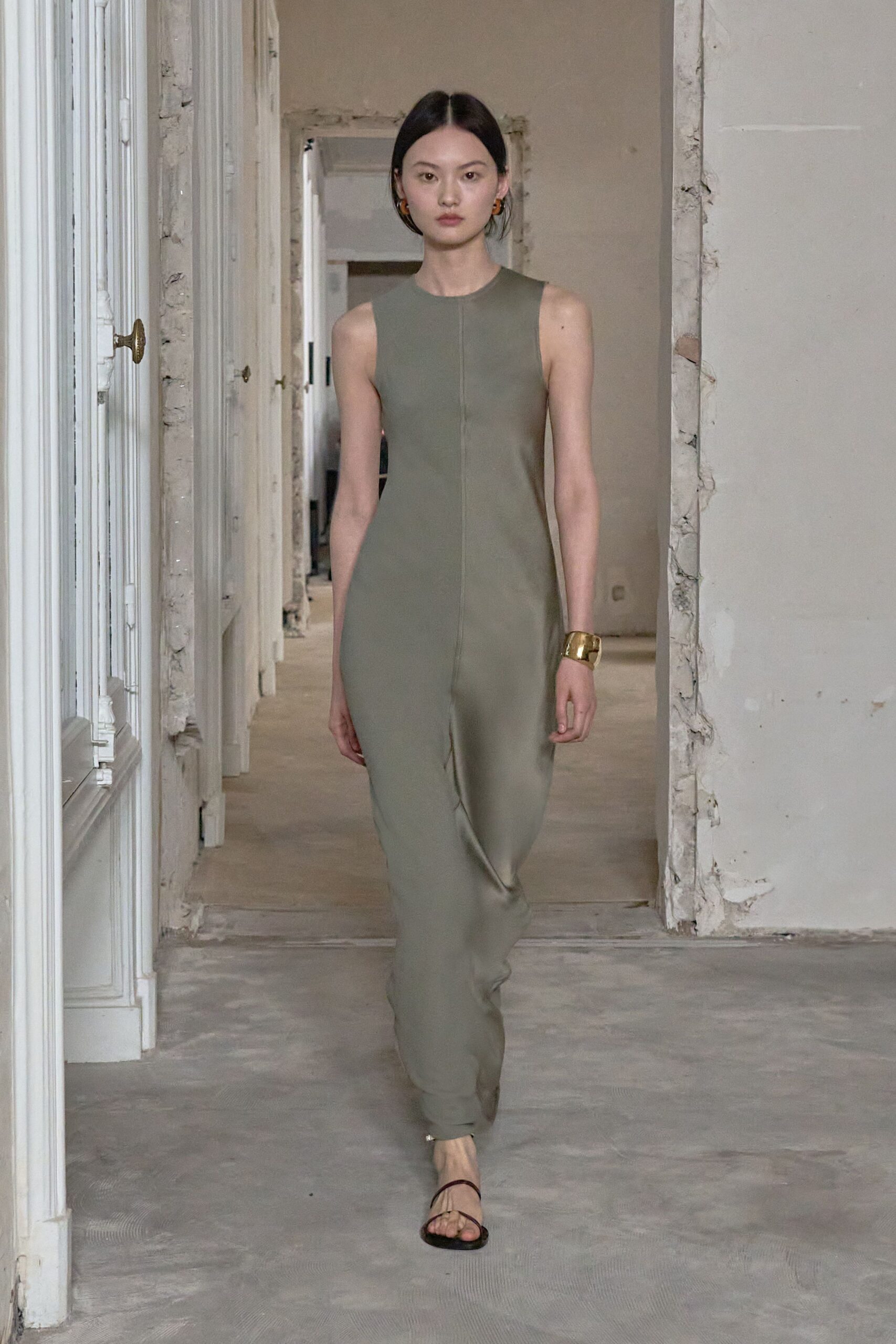 Sage column dress in matte crepe, split by a raw center seam. Wide hammered gold cuff. Amber resin hoops. Bare black thong sandals. Walking through an unfurnished apartment before the keys change hands.