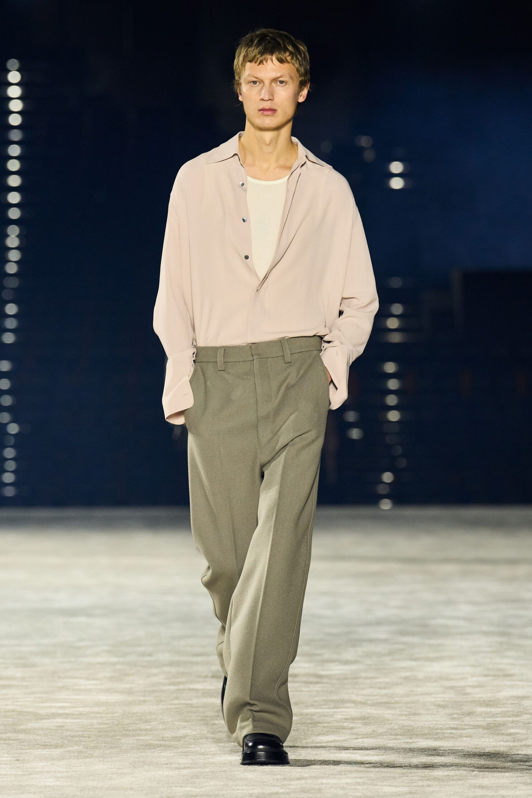 Blush fluid shirt, three dark buttons left casually undone. Cream ribbed tank peeking through the deep open placket. Sage twill trousers with a long clean crease and soft drape. Black square-toe shoes, barely breaking under the hem. Early checkout, hotel lobby still quiet except for the ice machine.