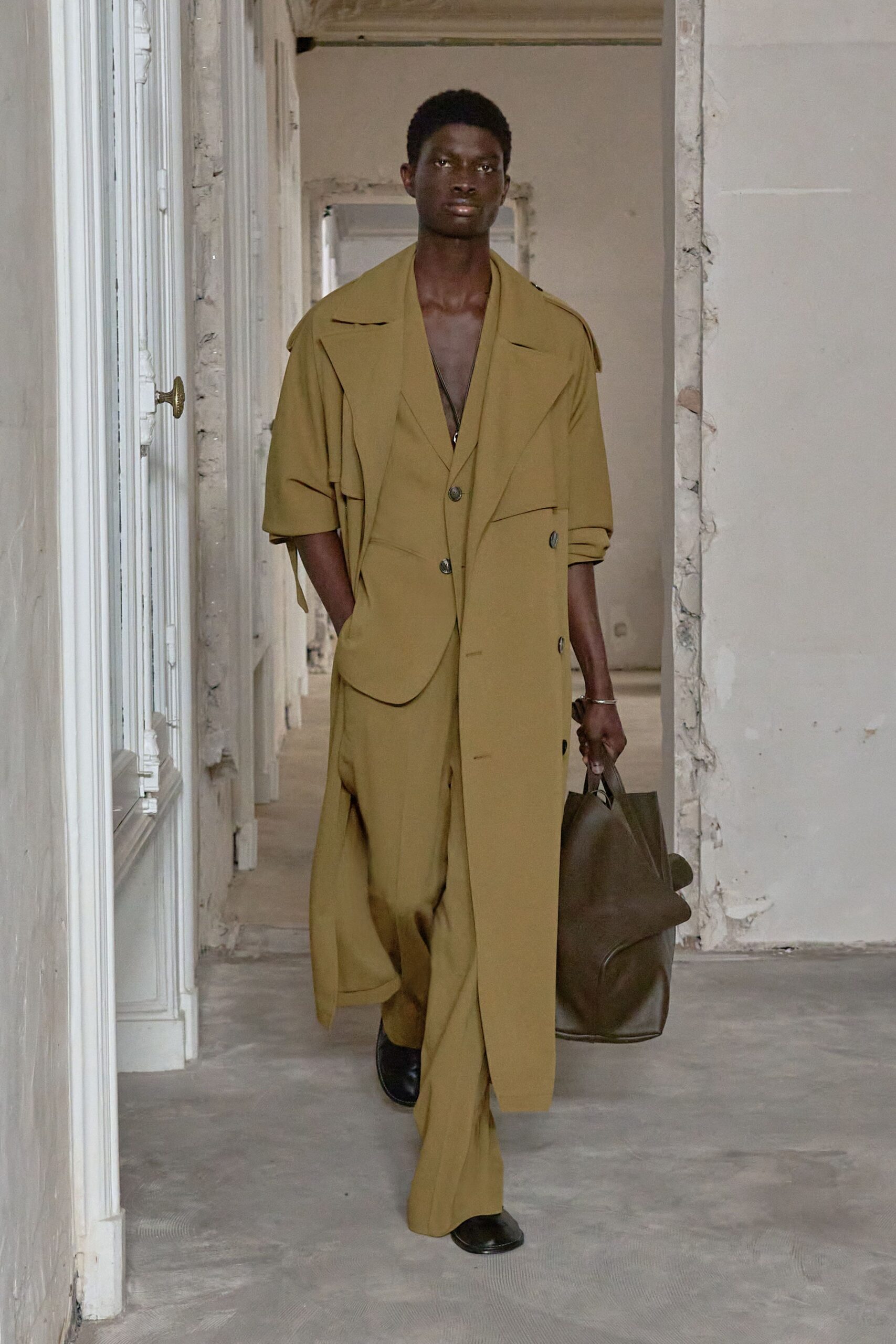 Olive draped coat with wide lapels and a hanging storm flap. Deep-V matching vest, one dark button catching at the waist. Loose olive trousers breaking long over the shoe. Oversized brown leather tote, soft sides collapsing inward. Black leather slip-ons. For crossing an empty hotel corridor before the meeting starts.