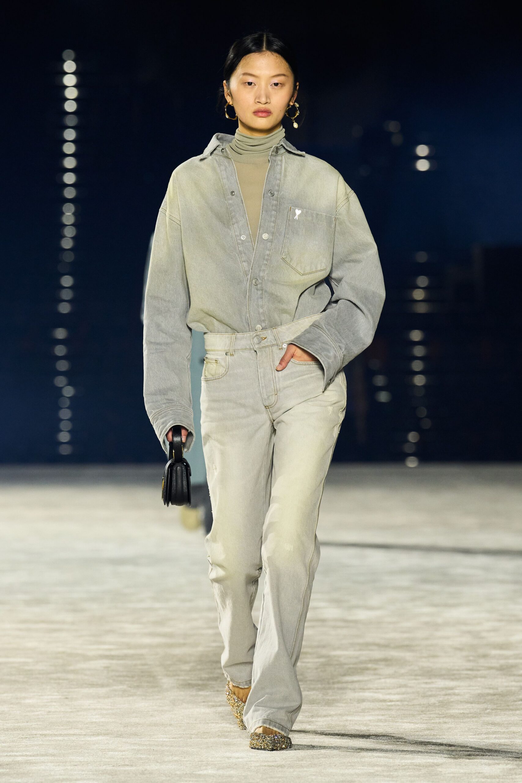 Taupe rib knit turtleneck, sheer through the chest. Washed stone denim shirt hanging loose, one chest pocket skewed low. Pale faded jeans with whiskering and a faintly abraded thigh. Small black top-handle bag. Gold glitter pumps. Walking across a marina promenade after the sun has dropped out.