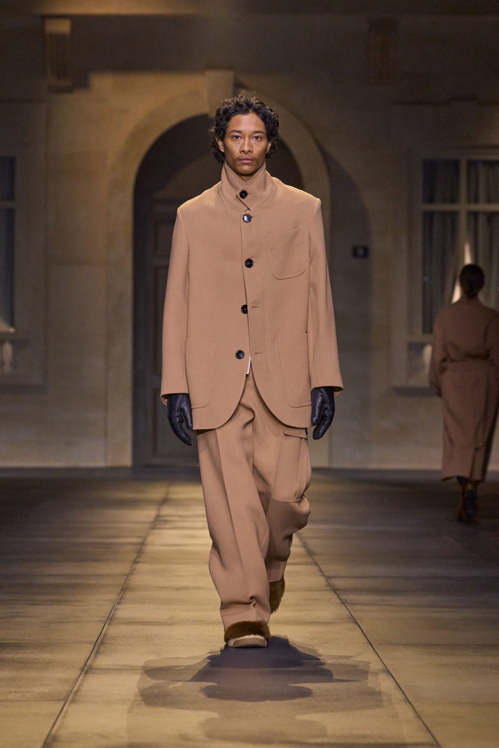 Camel wool jacket with a high funnel neck and glossy black buttons. Matching wide trousers with a curved utility pocket hanging off one thigh. Black leather gloves. Taupe slippers banded with thick brown fur at the vamp. Crossing a stone courtyard before sunrise, carrying the room's last warmth outside.