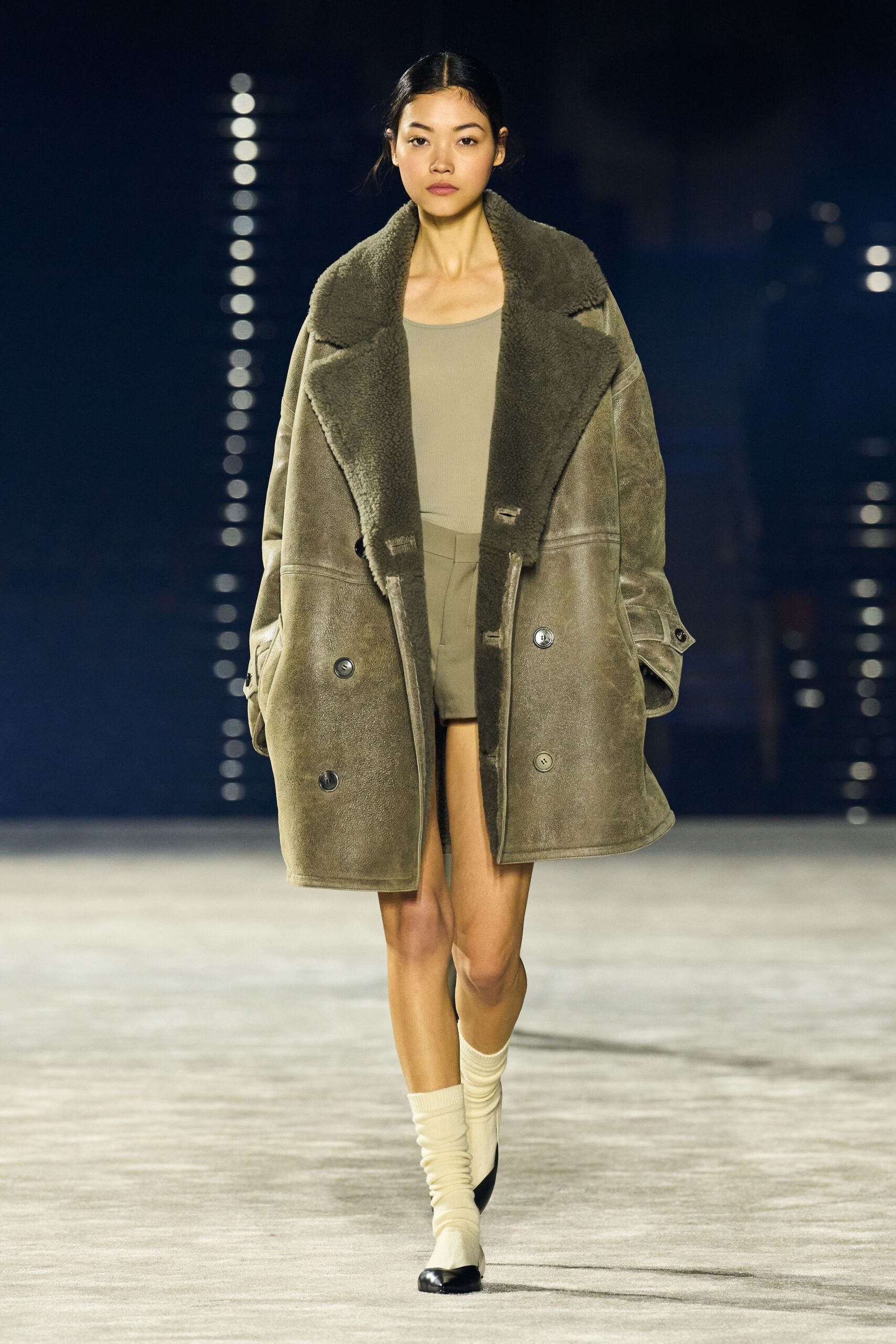 Oversized taupe shearling coat, broad lapels and matte horn buttons. Mushroom ribbed tank with a wide scooped neckline. High-waisted taupe shorts with a clean pressed front. Cream ribbed socks slouched at the ankle. Black cap-toe flats. First cold snap, crossing the parking deck before the office heat kicks on.