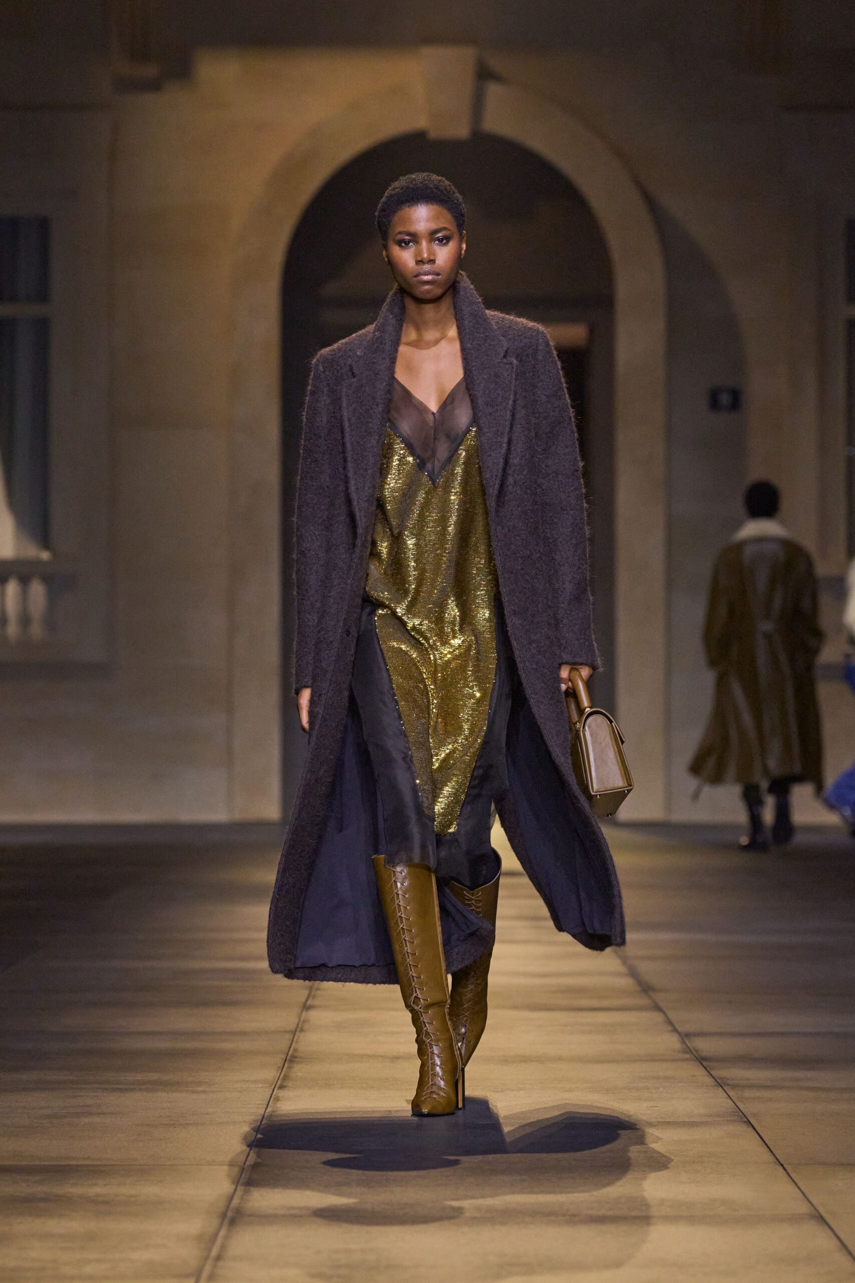 Long espresso brushed-wool coat with a thick shawl collar. Antique-gold sequined slip dress, plunged with smoky sheer mesh. Boxy tan top-handle bag with a rigid framed edge. Chestnut knee boots, corset-laced straight up the front. Crossing a hotel courtyard after midnight, key card warm in your palm.