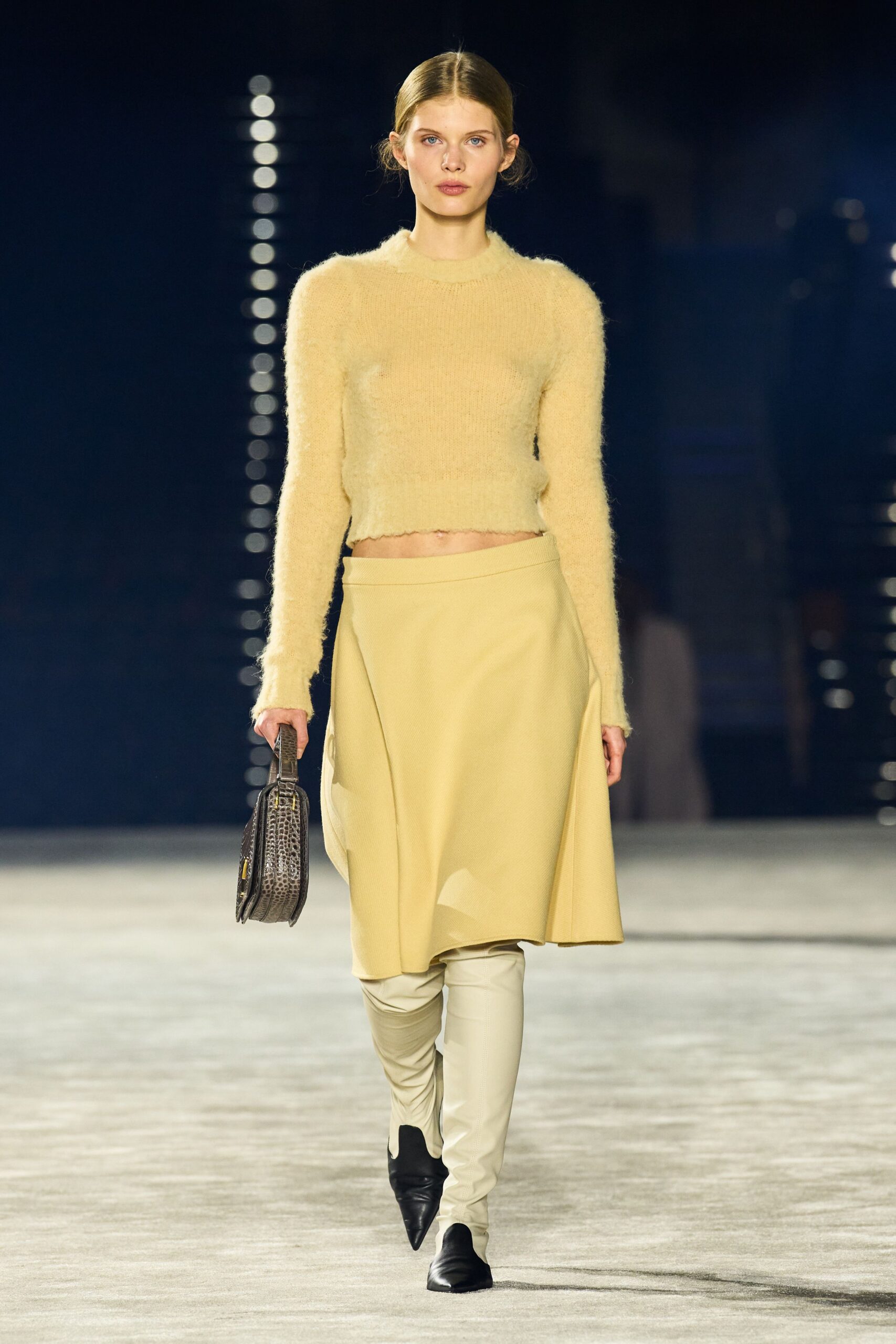 Buttercream fuzzy knit sweater, cropped with a lightly scalloped hem. Matching wrap skirt in dense crepe, one panel falling in a soft arc. Flat crocodile-embossed case bag with a rigid top handle. Cream thigh-high boots with black pointed toes. Early museum opening, carrying your notes before the lobby warms up.
