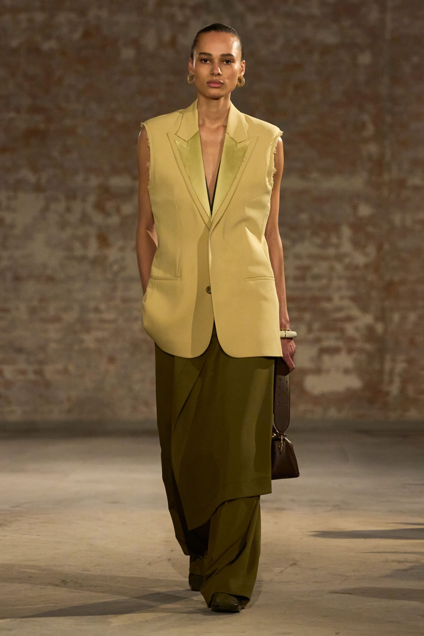 Buttercream tailored vest with raw-cut armholes and deep notched lapels. Olive wrap skirt, one sharp panel slanting across the front. Loose olive trousers pooling under the skirt layer. Small twisted gold hoops. Dark brown shoulder bag with a wide punched strap. Crossing a museum courtyard before the heat leaves the stone.