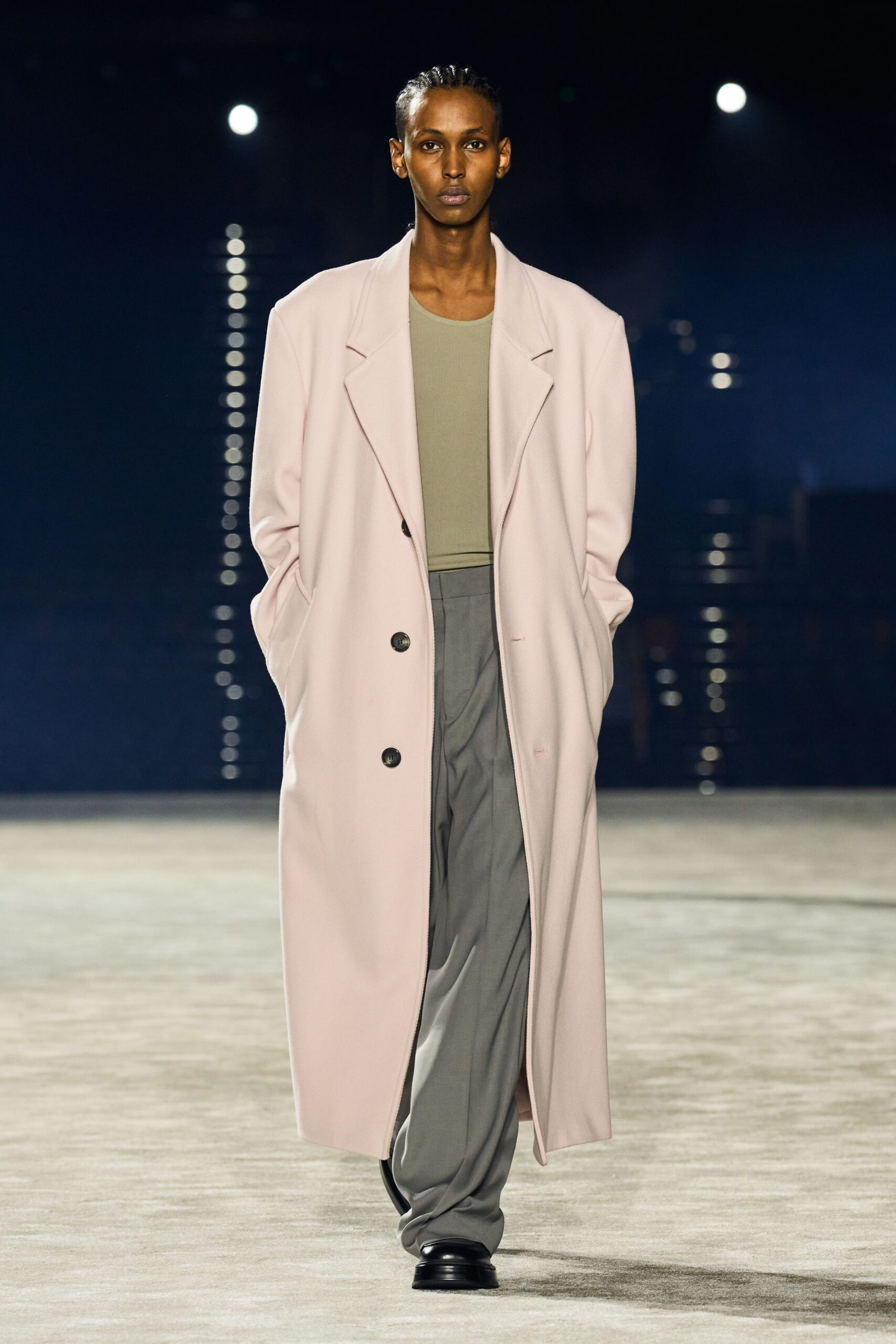 Pale blush overcoat in dense twill, dropped shoulders and dark horn buttons. Sage ribbed tank cut close at the collarbone. Slate trousers with a soft front drape pooling over the shoes. Black leather square toes. First train out, station concourse still smelling of detergent and rain.