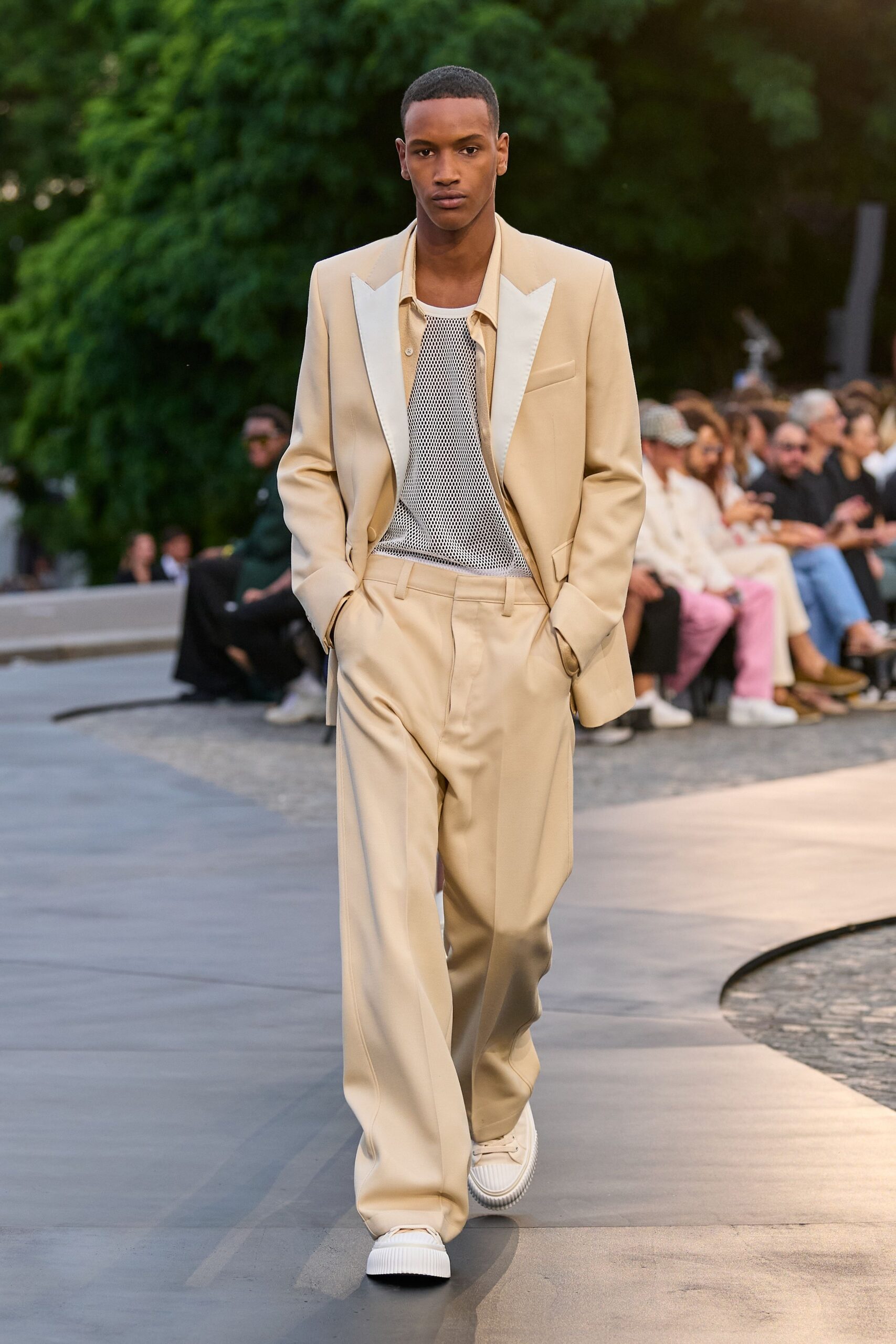 Sand tailoring with broad ivory satin peak lapels. Pale camp-collar shirt left open under the jacket. White open-mesh tank laid over a solid underlayer. Matching wide-leg trousers with a soft break over the shoe. White ribbed-sole sneakers. Crossing a hotel courtyard just before the wedding guests sit down.