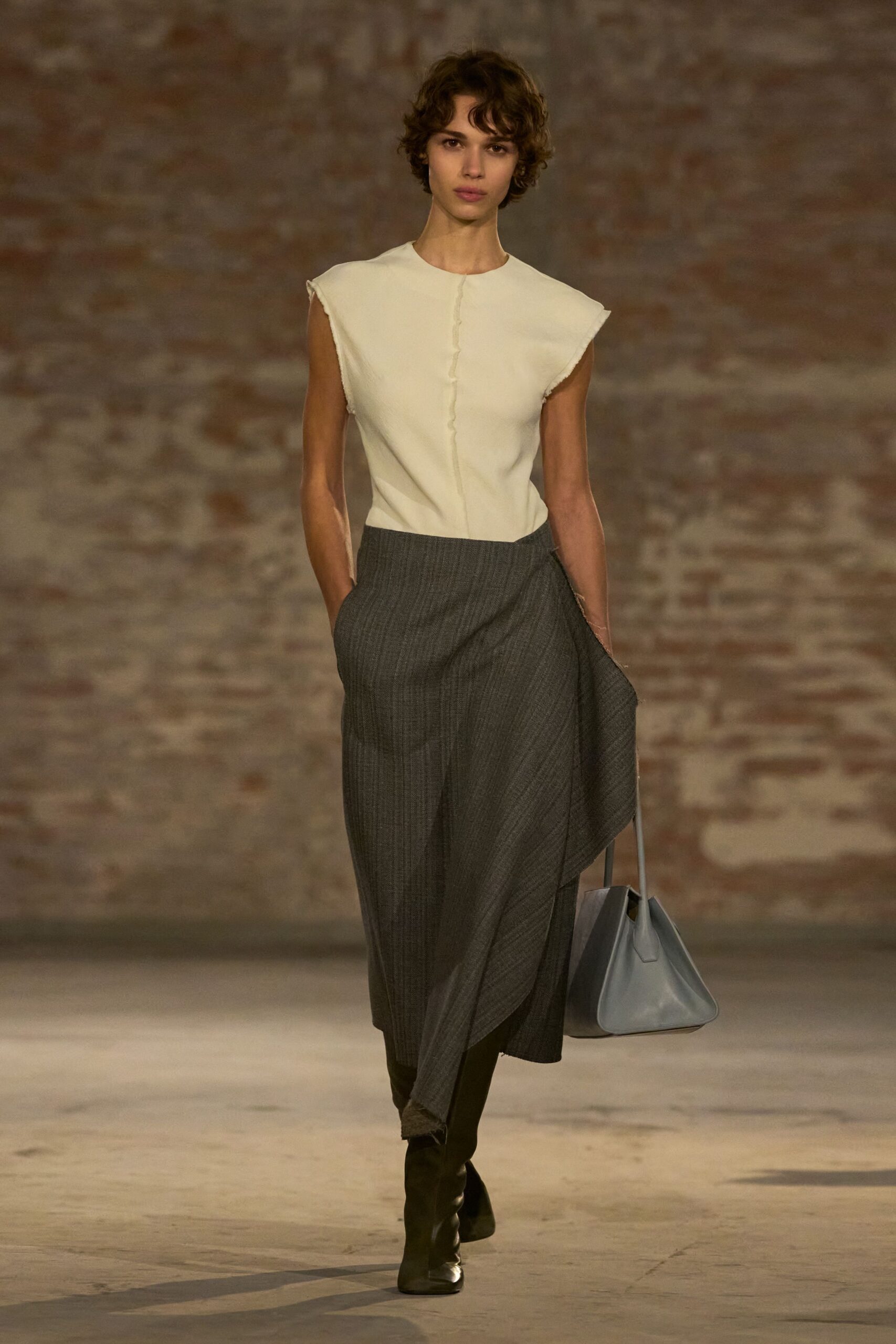 Cream sleeveless knit top with raw armholes and a rippled center seam. Charcoal pinstripe wrap skirt, one panel hanging loose with a frayed edge. Slate leather tote with rigid trapezoid sides. Black knee boots with square toes. For crossing a museum courtyard in weather that cannot decide.