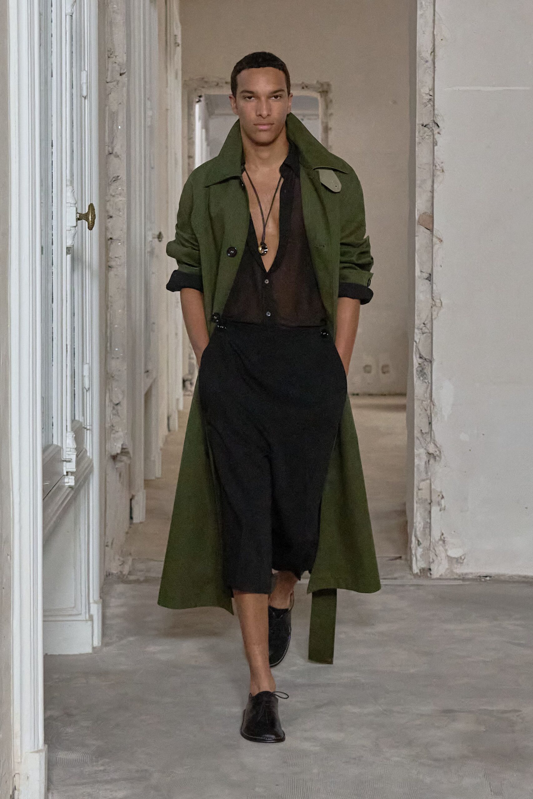 Moss green coat with a high stand collar and hanging belt end. Sheer black shirt worn open to the navel, tiny buttons catching light. Black cord pendant necklace. Black cropped trousers, loose through the thigh. Black leather lace-up shoes. Crossing a cold villa corridor before anyone opens the shutters.